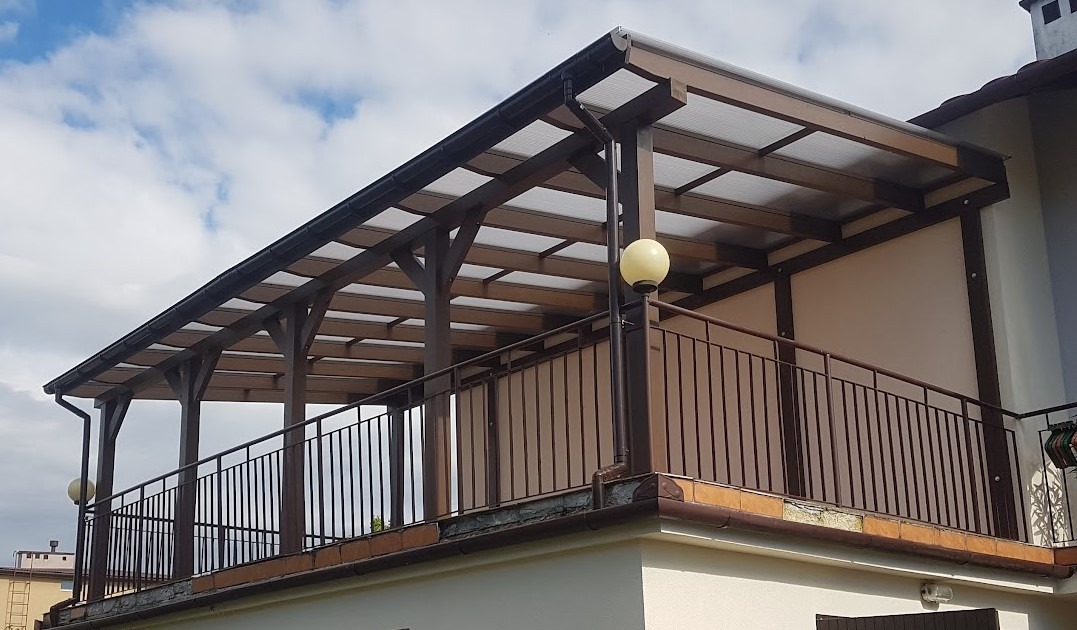 Zadaszona, drewniana konstrukcja pergoli z przezroczystym dachem, zamontowana na balkonie budynku, z widocznymi drewnianymi słupami i metalową balustradą w kolorze brązowym.