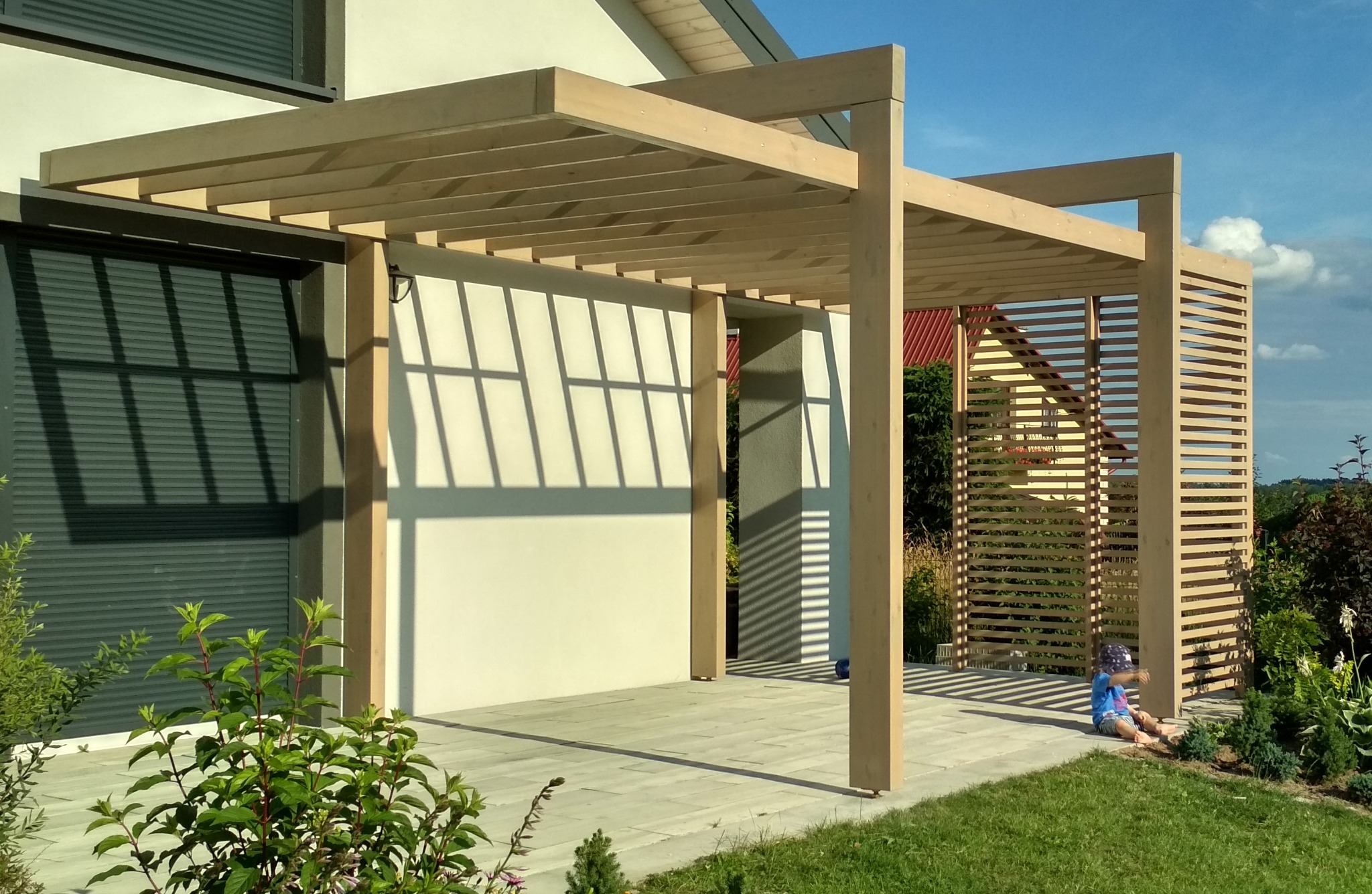 Surowa, jasna konstrukcja z drewna tworząca zadaszenie tarasu, z elementami ażurowymi z boku, rzucająca cień na kamienną posadzkę; w tle dom z szarymi roletami i błękitne niebo, na pierwszym planie...