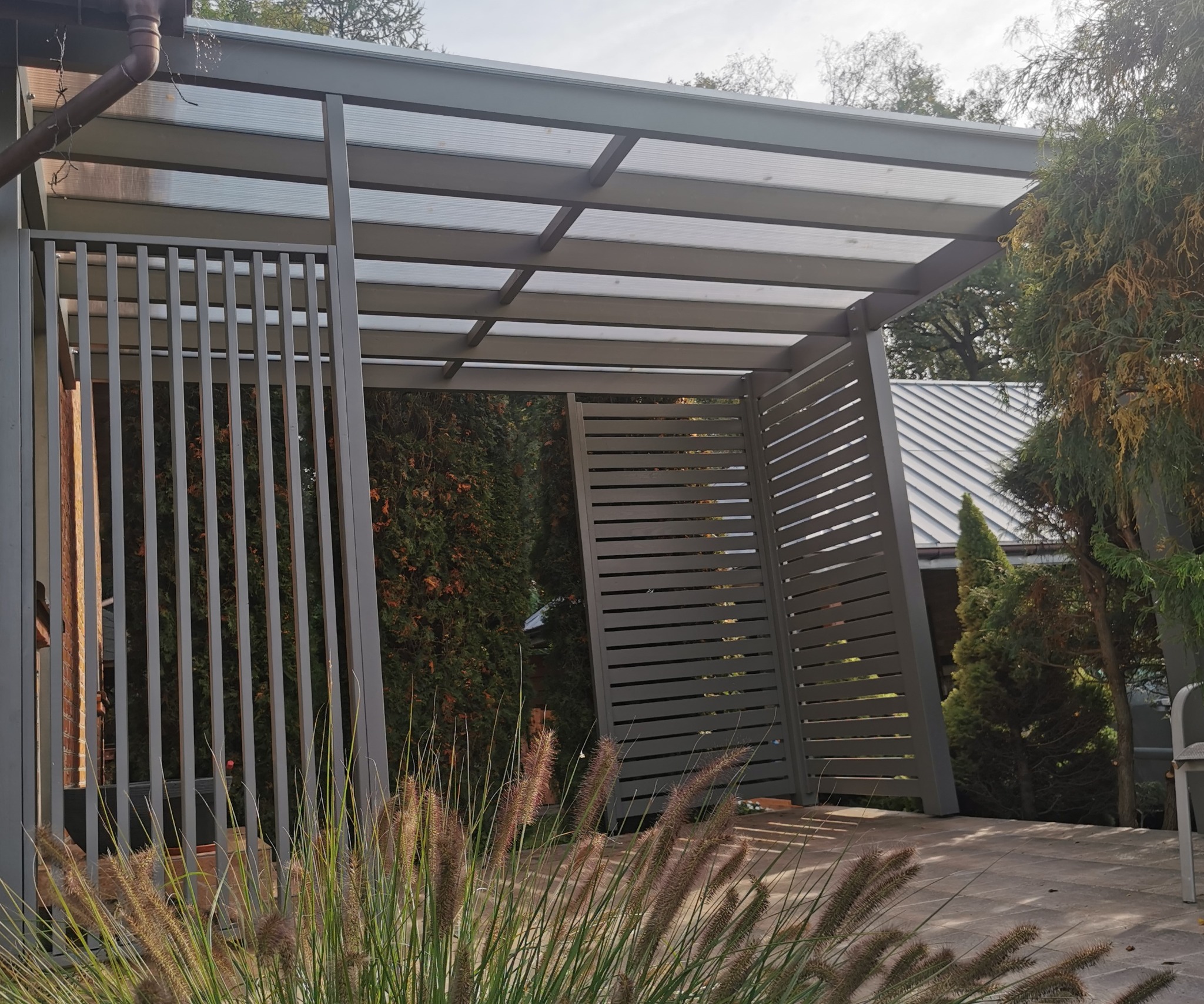 Nowoczesna, szara konstrukcja z zadaszeniem poliwęglanowym i ażurowymi ściankami, otoczona zielenią, z widocznymi trawami na pierwszym planie.