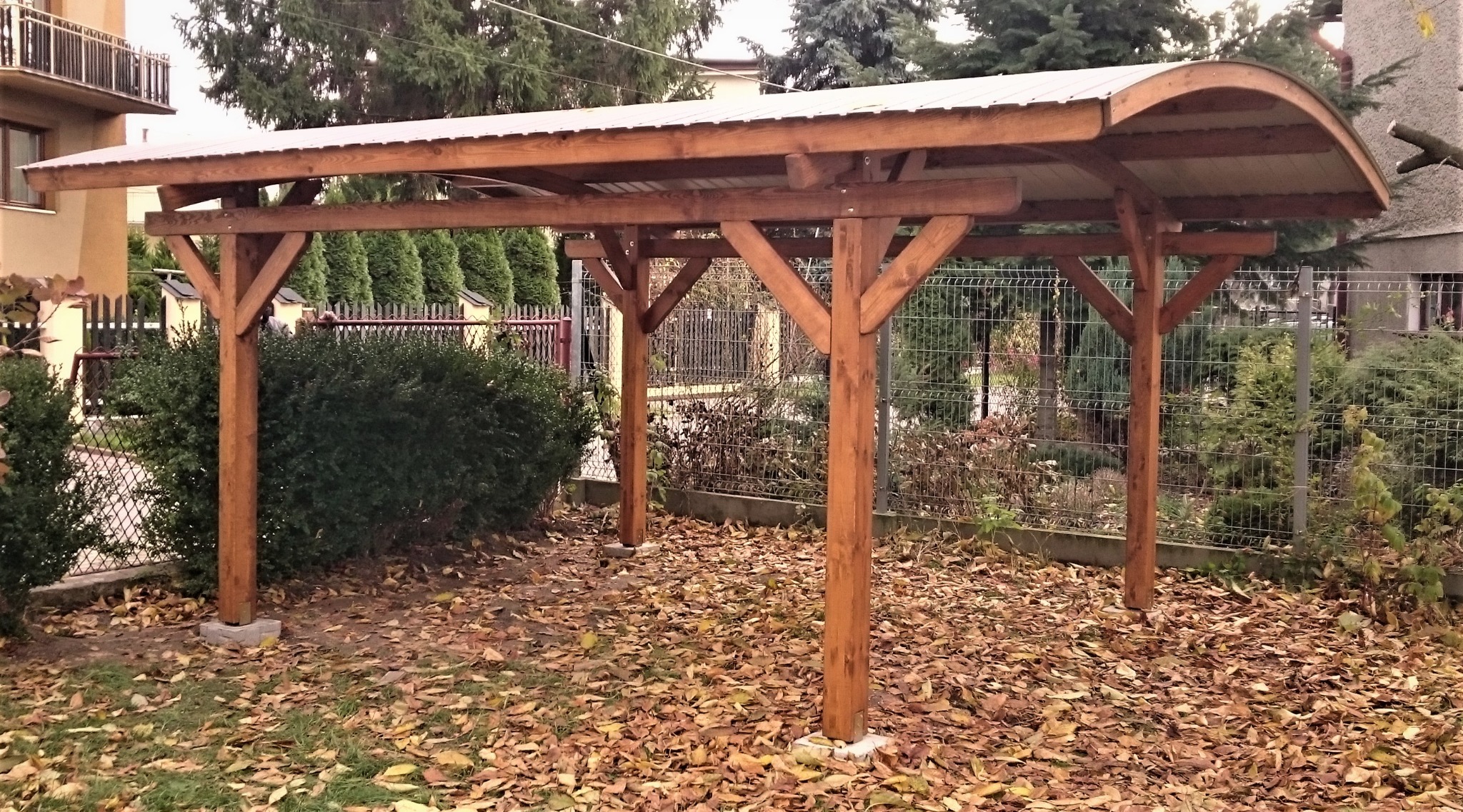 Zadaszona konstrukcja drewniana z metalowym dachem, wsparta na betonowych fundamentach, otoczona opadłymi liśćmi i ogrodzeniem z siatki.