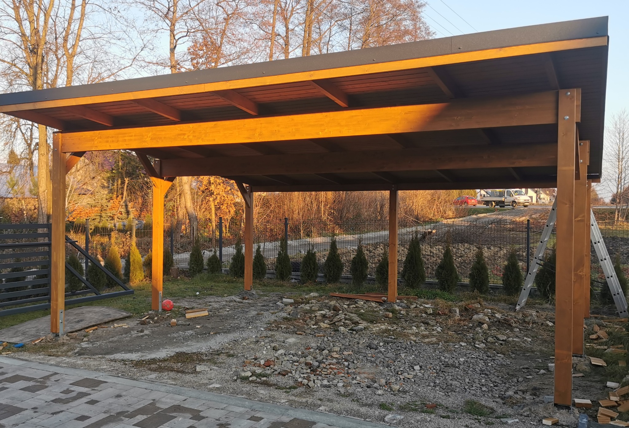 Drewniana konstrukcja z płaskim dachem w trakcie budowy, widoczne słupy nośne, fragment dachu, gruz na podłożu, drabina oparta o konstrukcję, w tle żywopłot z iglaków.