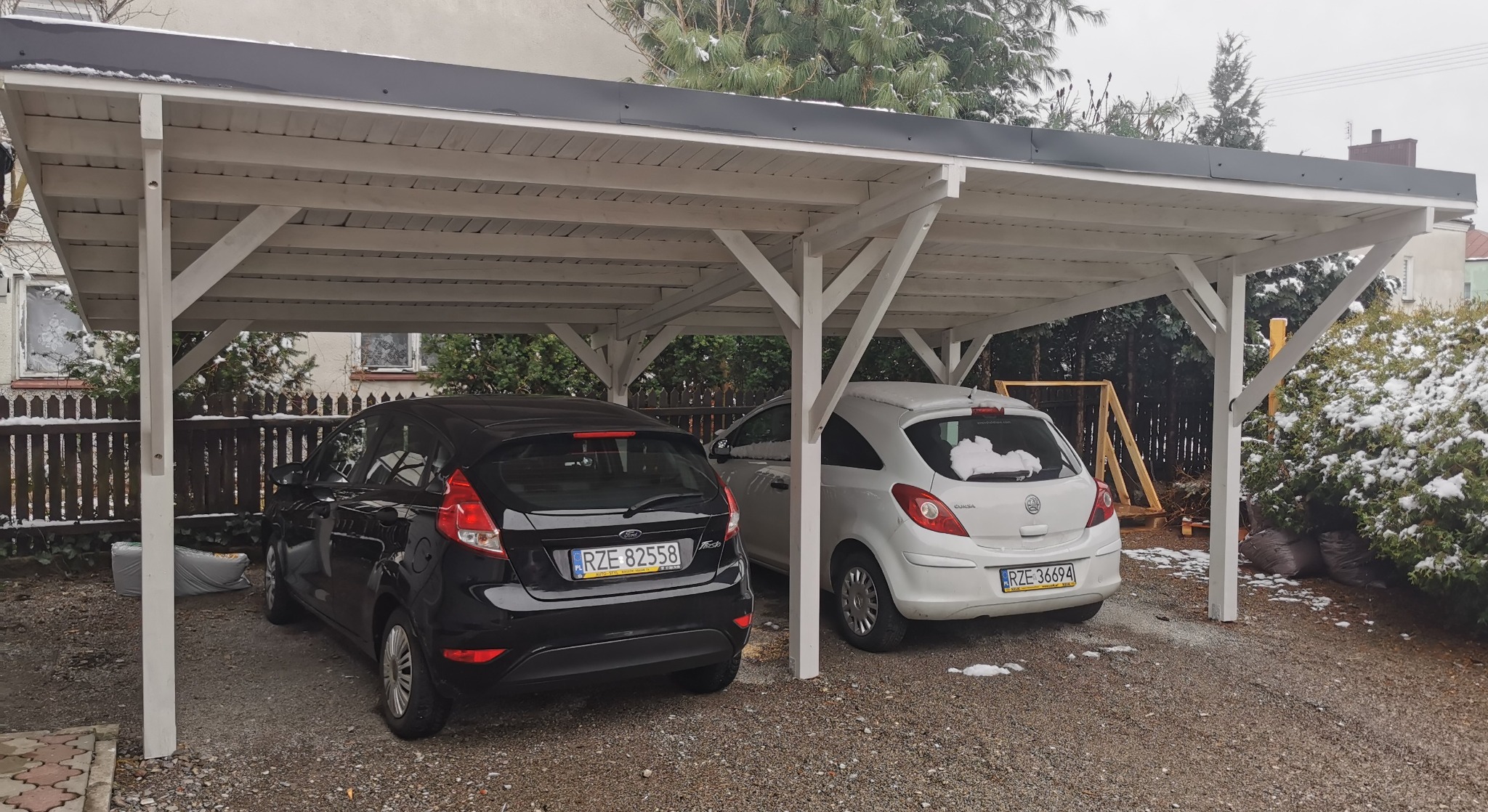 Biała, drewniana konstrukcja z płaskim dachem, służąca jako wiata garażowa dla dwóch samochodów w Sokołowie Małopolskim, z lekkim opadem śniegu.