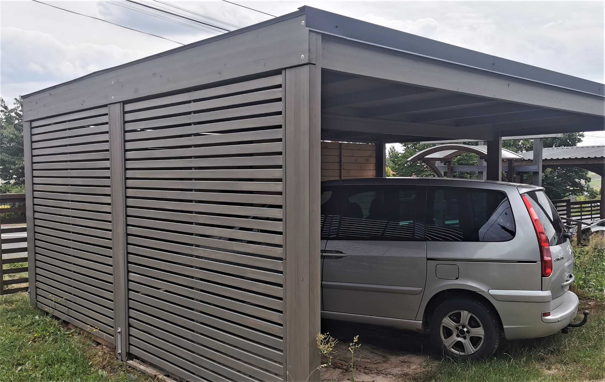 Szara konstrukcja z drewnianych listew, pełniąca funkcję zadaszonego miejsca parkingowego, z zaparkowanym srebrnym minivanem w środku.