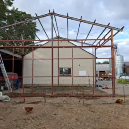 Stalowa konstrukcja hali w trakcie budowy na terenie wiejskim, widoczne kury grzebiące w ziemi, budynek z cegły w tle.