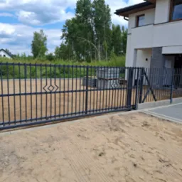 Nowoczesna, przesuwna brama wjazdowa z metalowymi prętami i ozdobnym rombem, zamontowana przed nowym domem z jasną elewacją, widoczne kostki brukowe i piasek.