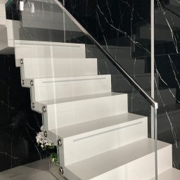 METAL-CHROM - Nowoczesne, białe schody z przezroczystą balustradą szklaną i chromowanymi detalami, na tle ściany z czarnego marmuru z białymi żyłkami. Dekoracja kwiatowa u podstawy schodów.