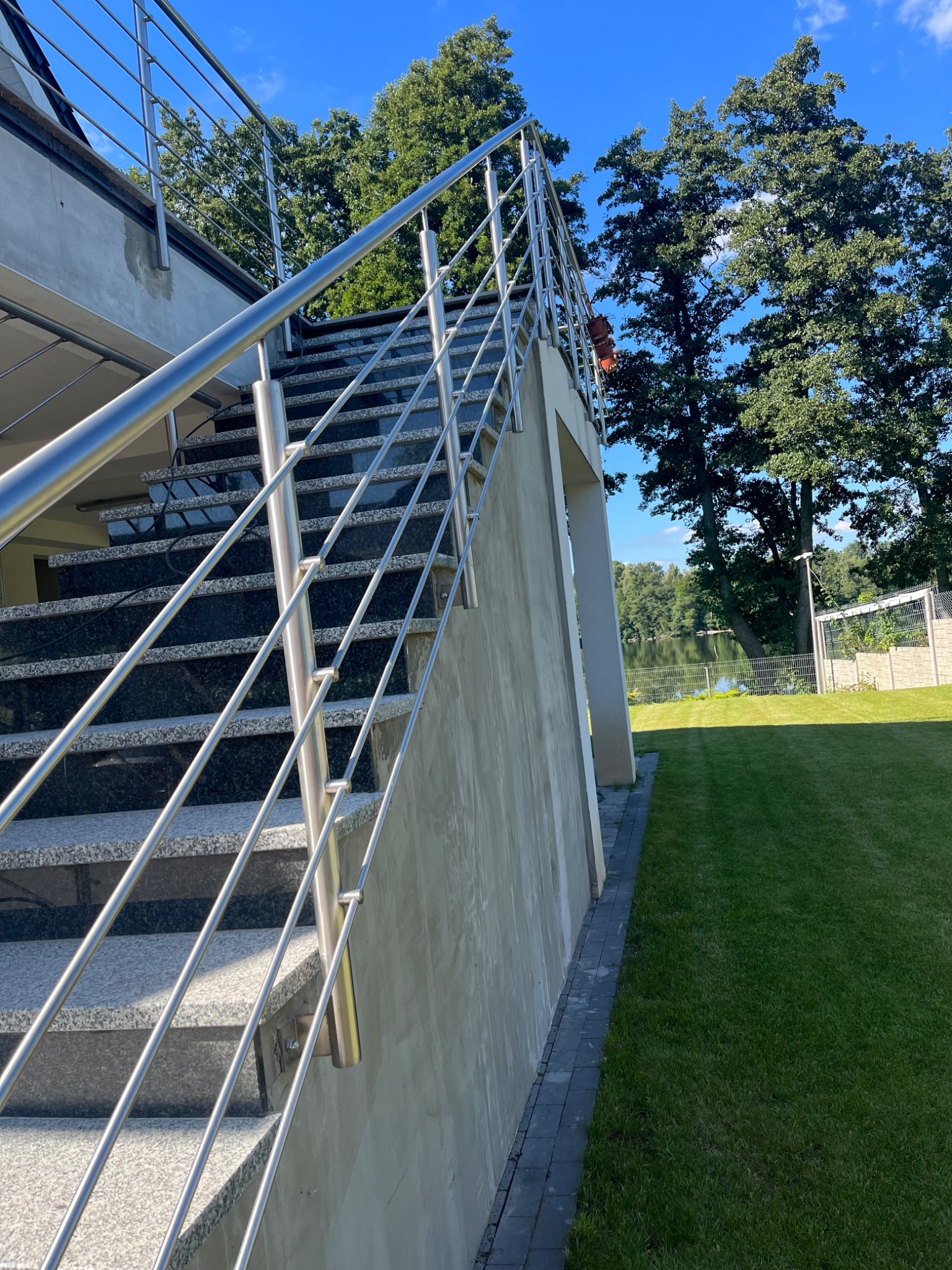 Zewnętrzne schody z szarego granitu z nowoczesną balustradą ze stali nierdzewnej w Wałczu, widok z dołu na tle zielonego trawnika i jeziora.