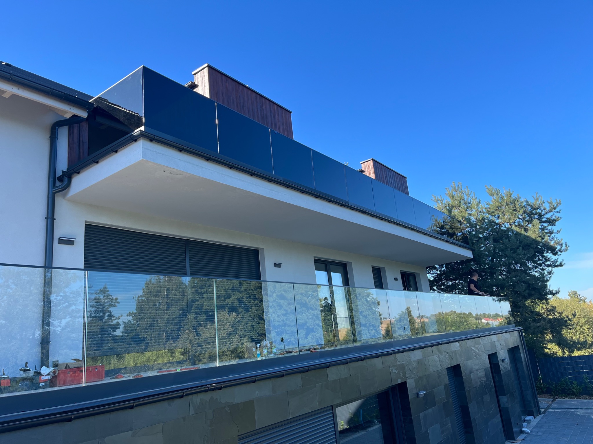 Nowoczesny budynek z balkonem i szklaną balustradą, widok z dołu na błękitne niebo, z widocznymi narzędziami i materiałami budowlanymi na balkonie.