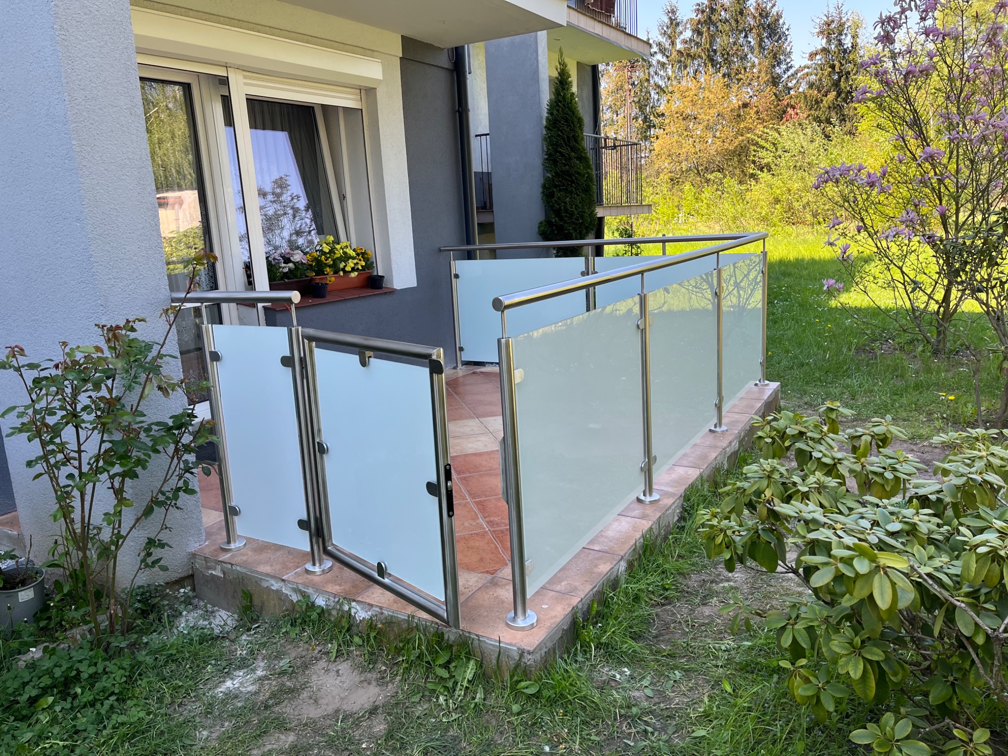 Zewnętrzna balustrada ze szkła mlecznego i stali nierdzewnej, z otwartymi drzwiczkami, otoczona zielenią i kwiatami, przylegająca do tarasu z terakotową podłogą.
