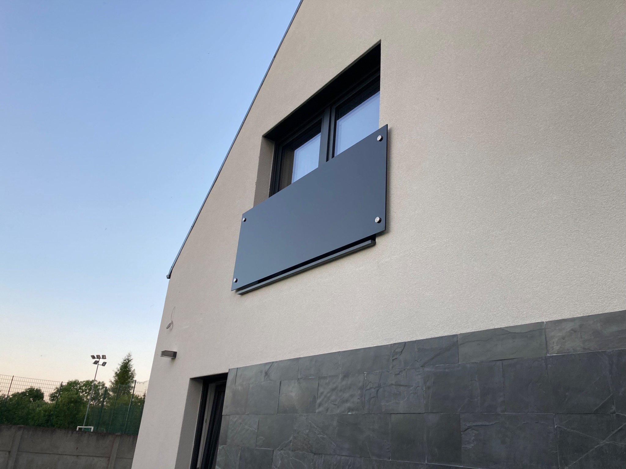 Minimalistyczna balustrada balkonowa z ciemnoszarej płyty przymocowana do elewacji budynku, widok z dołu na tle błękitnego nieba.