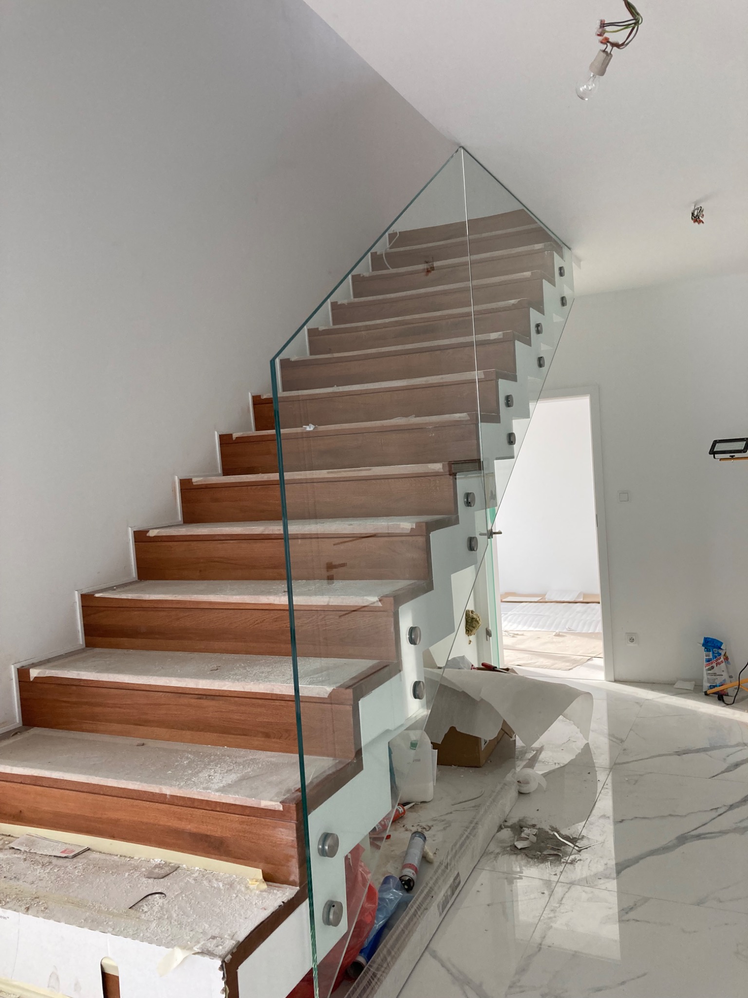 Wnętrze w trakcie wykańczania z drewnianymi schodami i szklaną balustradą, zabezpieczone folią malarską, widoczne niedokończone oświetlenie sufitowe i marmurowe płytki podłogowe.
