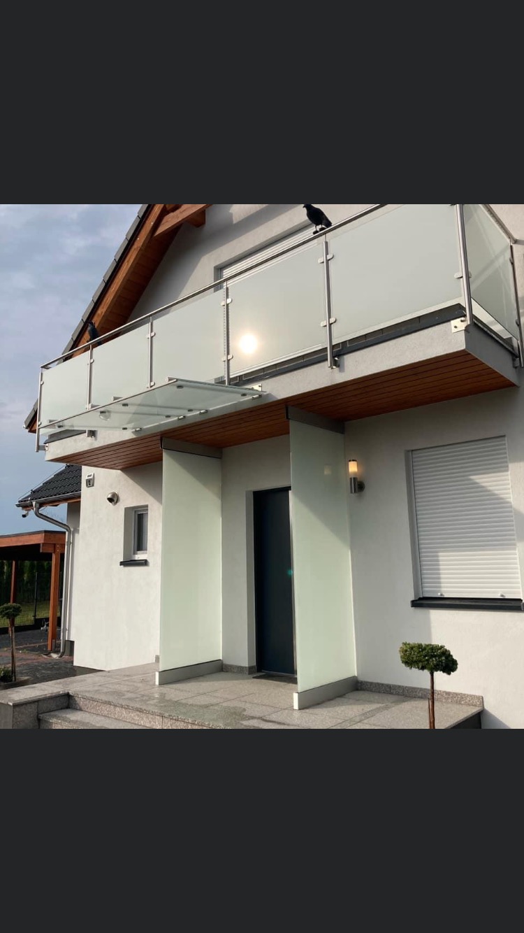 Nowoczesny dom z balkonem zabezpieczonym szkłem i stalą nierdzewną, z widocznymi dwoma krukami na balustradzie, oraz szklanym zadaszeniem nad wejściem.
