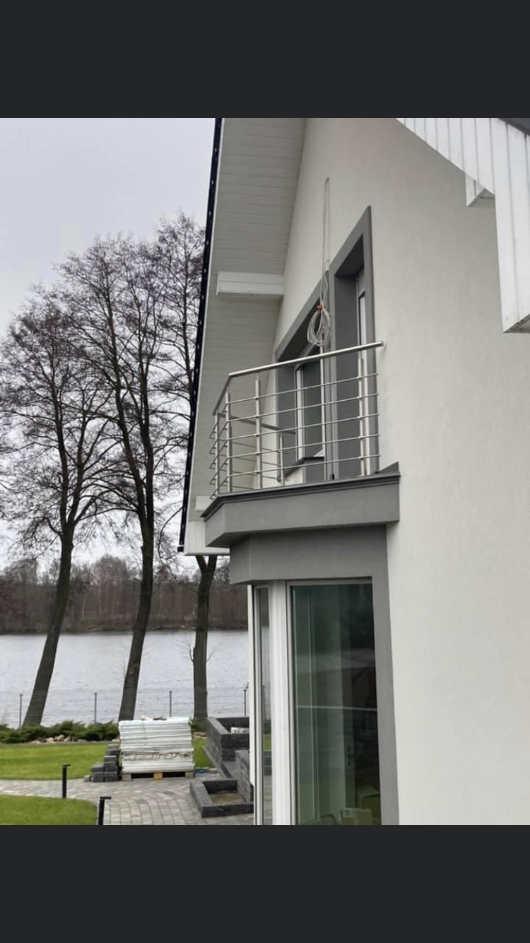 Nowoczesny balkon z prostą, stalową balustradą na tle jeziora i nagich drzew, widoczne elementy elewacji domu i prace brukarskie w ogrodzie.