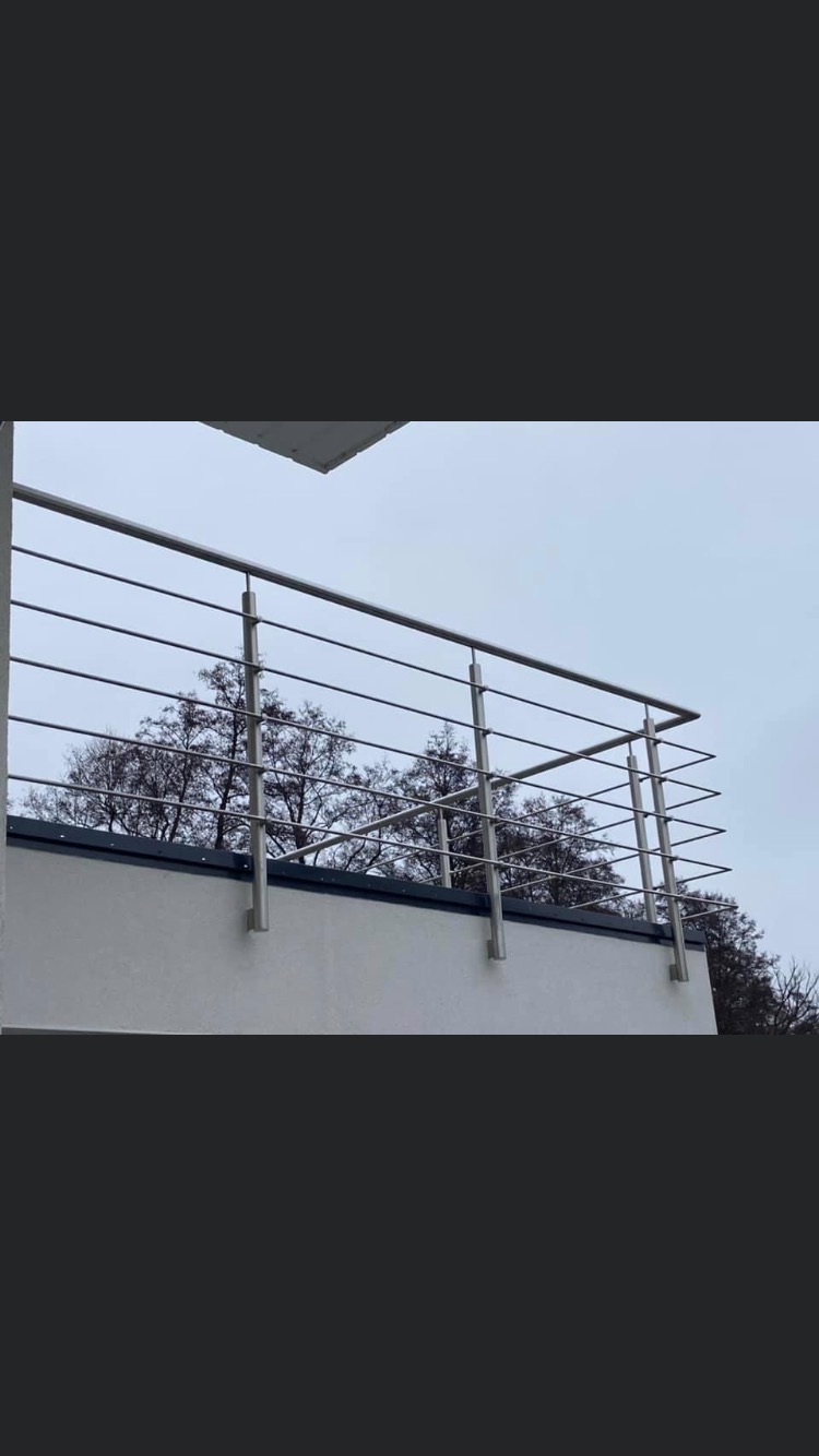 Minimalistyczna balustrada balkonowa ze stali nierdzewnej z prostymi, poziomymi prętami, zamontowana na białej ścianie budynku, z widokiem na drzewa bez liści na tle pochmurnego nieba.