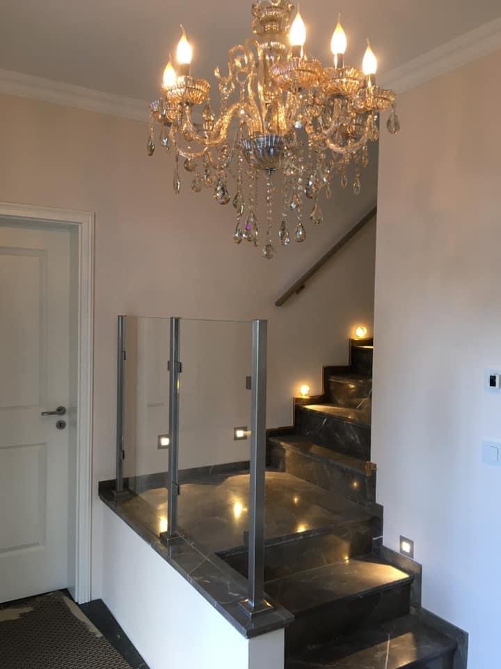 Nowoczesne schody z ciemnego marmuru z podświetleniem stopni i szklaną balustradą na metalowych słupkach, oświetlone kryształowym żyrandolem.