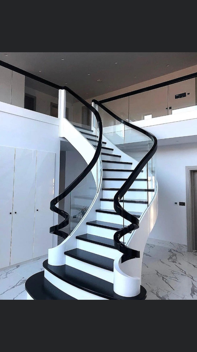 Eleganckie, spiralne schody z czarnymi stopniami i białą konstrukcją, otoczone szklaną balustradą z czarnymi poręczami, w przestronnym, nowoczesnym wnętrzu z marmurową podłogą.