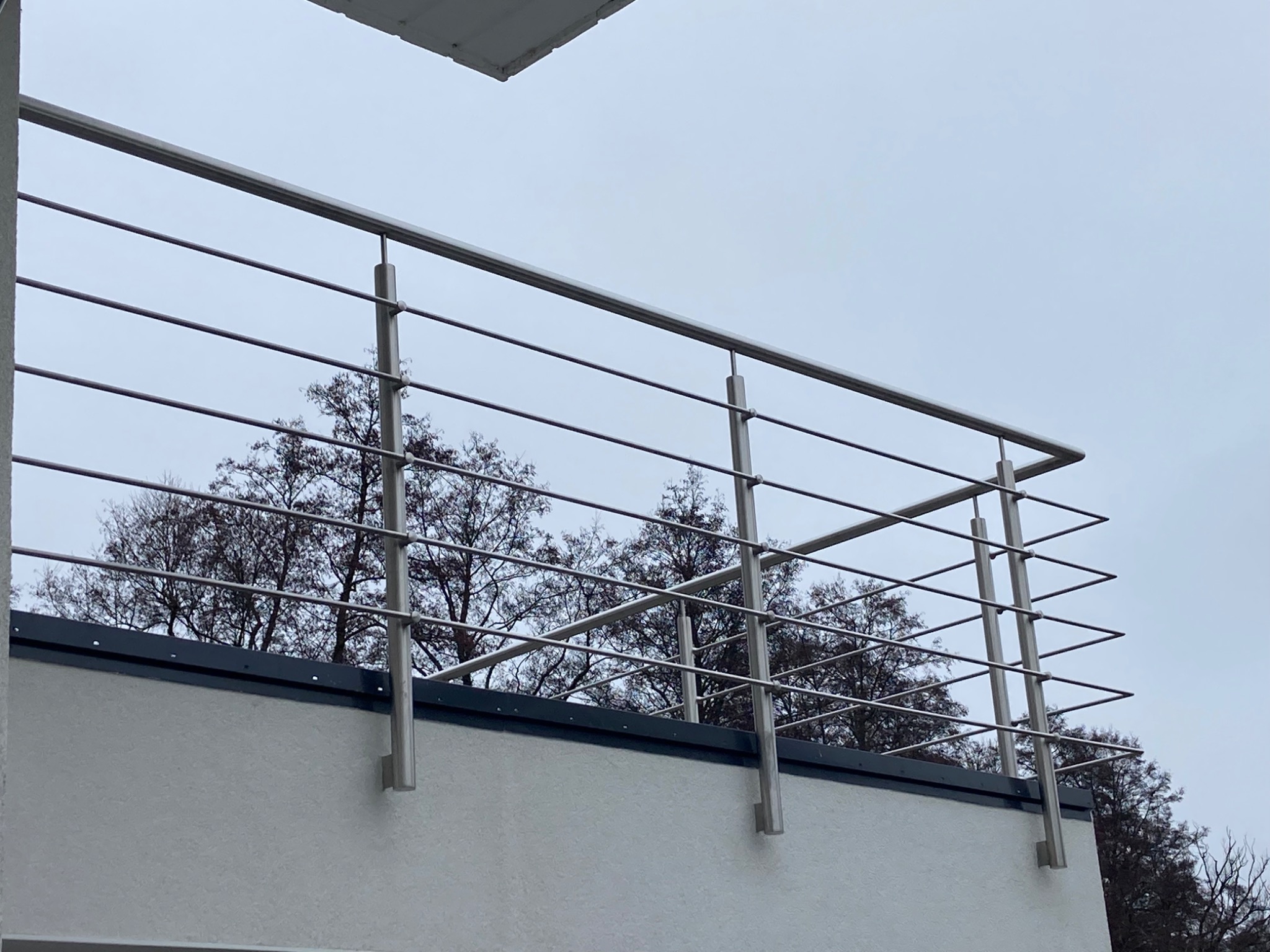 Nowoczesna balustrada nierdzewna z poziomymi prętami na balkonie, widok z dołu, na tle drzew bez liści i pochmurnego nieba.