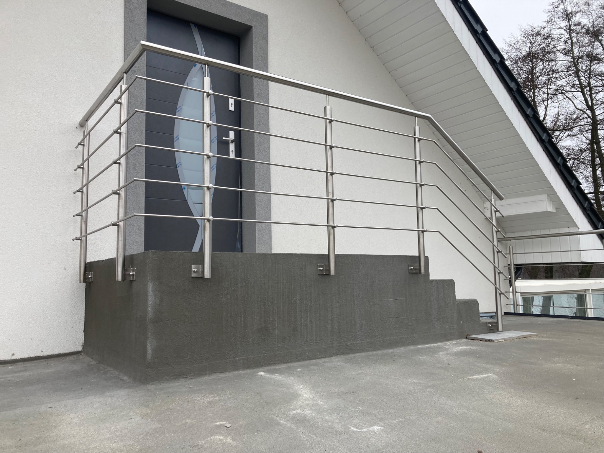 Zewnętrzne schody z surowego betonu z nowoczesną balustradą ze stali nierdzewnej, mocowaną na kwadratowych wspornikach, prowadzące do drzwi wejściowych z dekoracyjnym przeszkleniem.