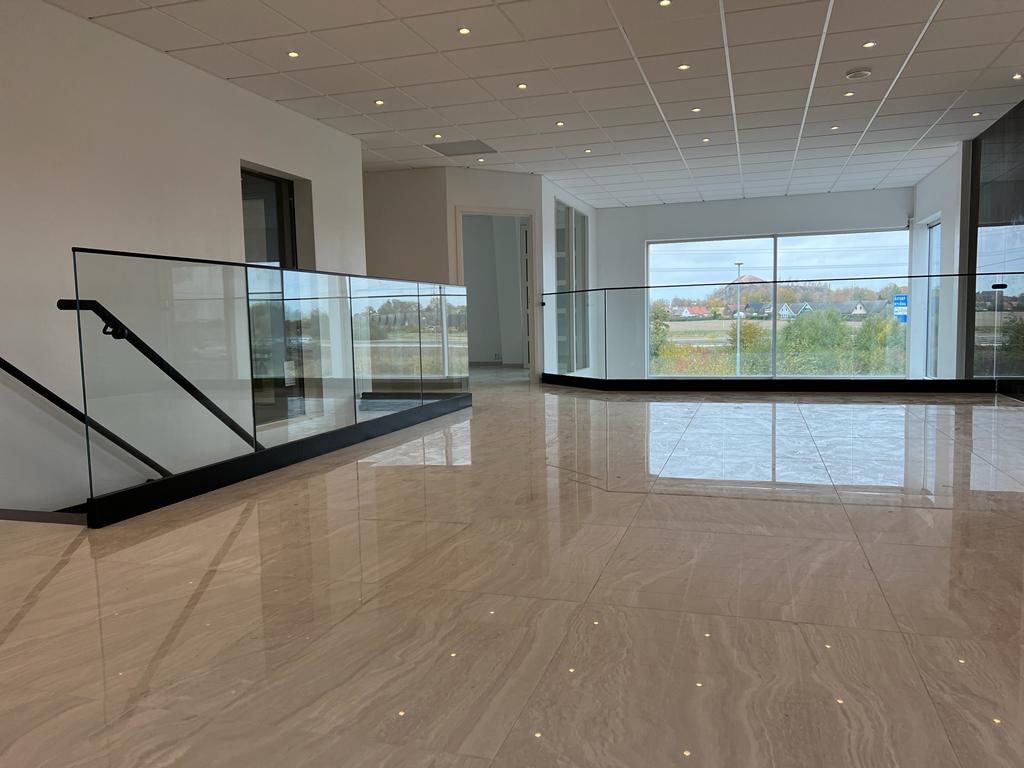 Wnętrze nowoczesnego budynku z błyszczącą marmurową podłogą, przeszkloną balustradą z czarnymi elementami oraz dużym oknem z widokiem na krajobraz.