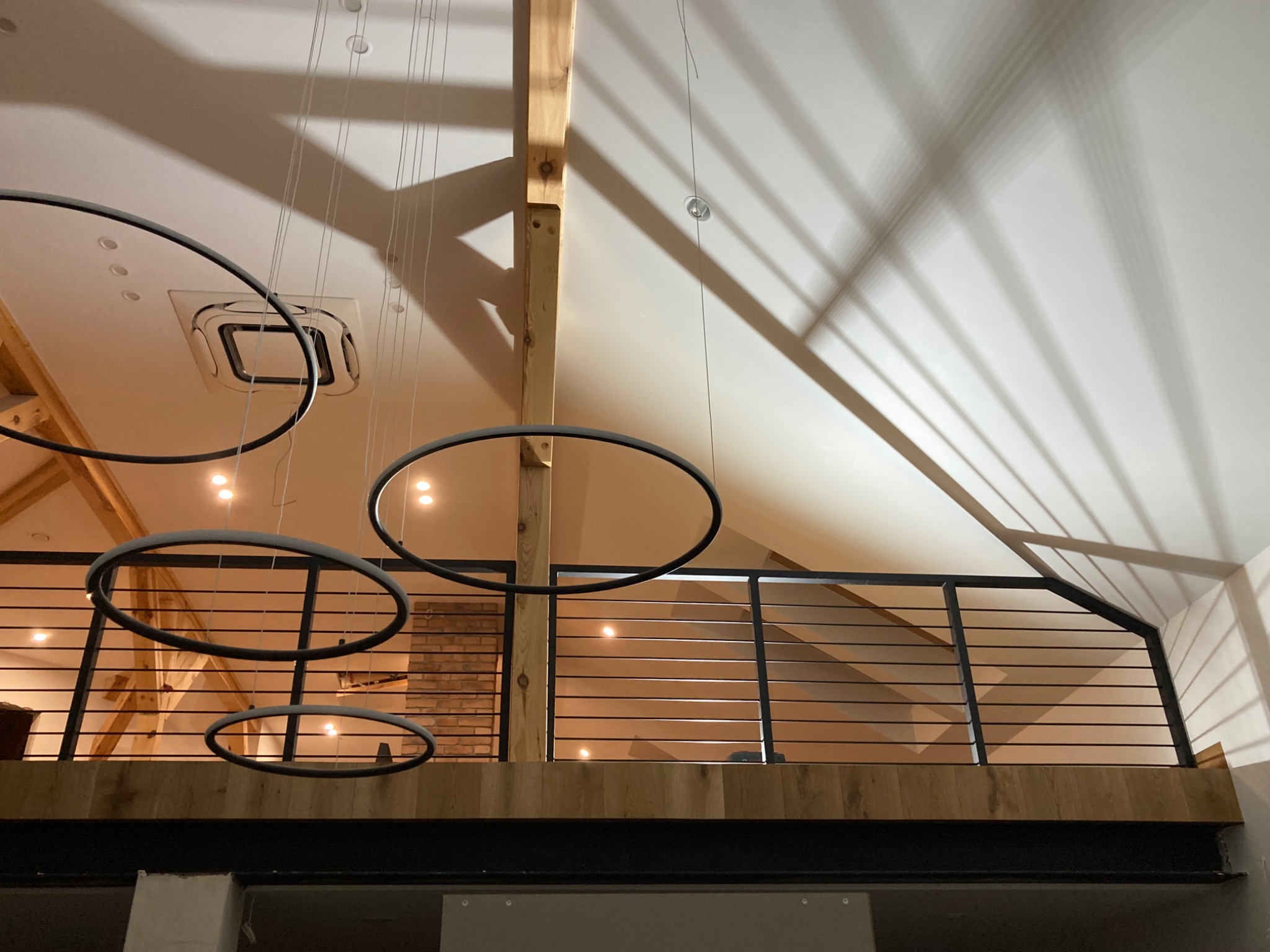 Wnętrze z antresolą zabezpieczoną poziomą, metalową balustradą, widoczne drewniane elementy konstrukcyjne, okrągłe lampy wiszące i cień konstrukcji dachu na suficie.