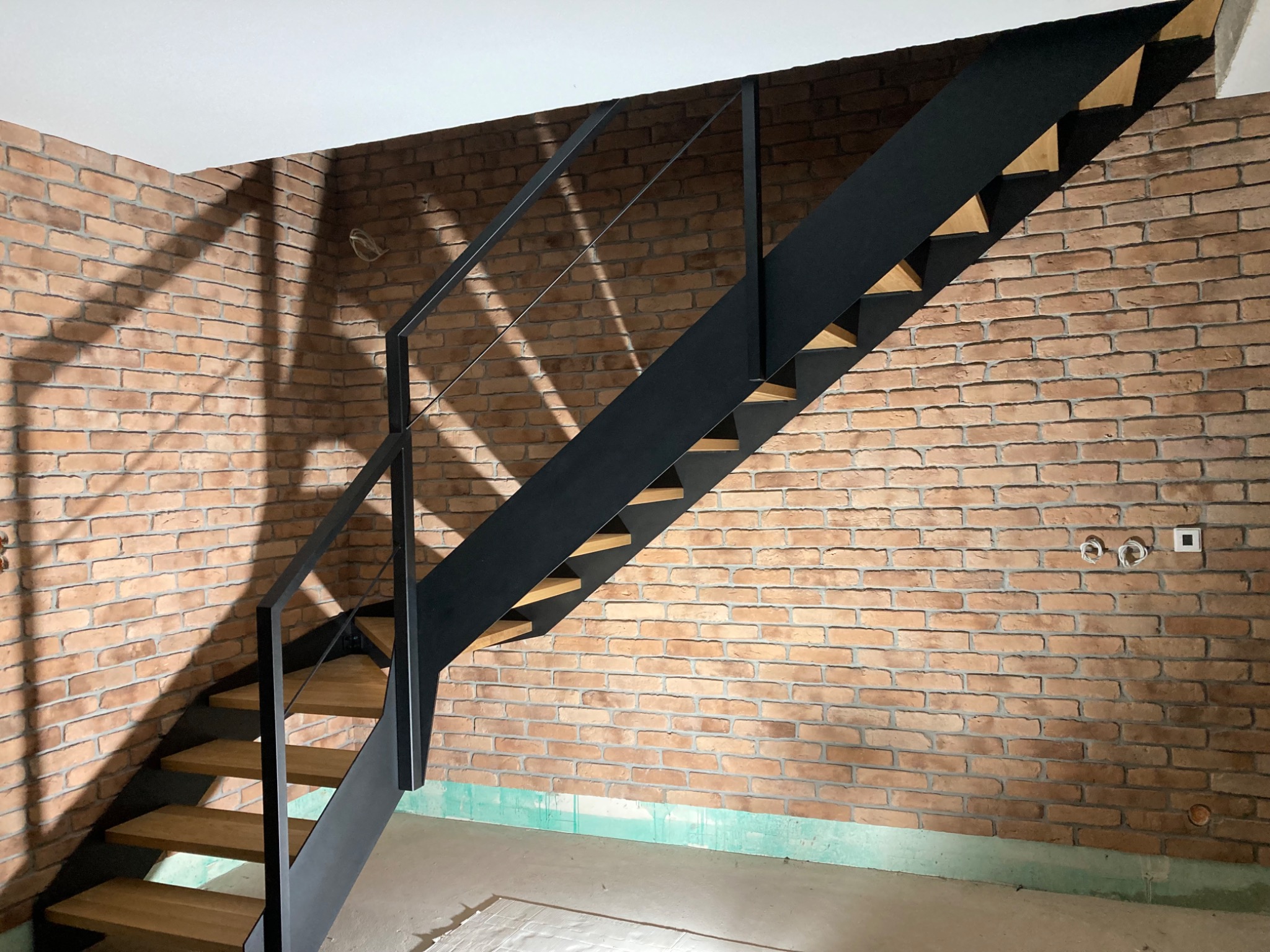 Nowoczesne schody metalowe z drewnianymi stopniami na tle ściany z cegły, widoczne kable elektryczne i niedokończone wykończenie podłogi.