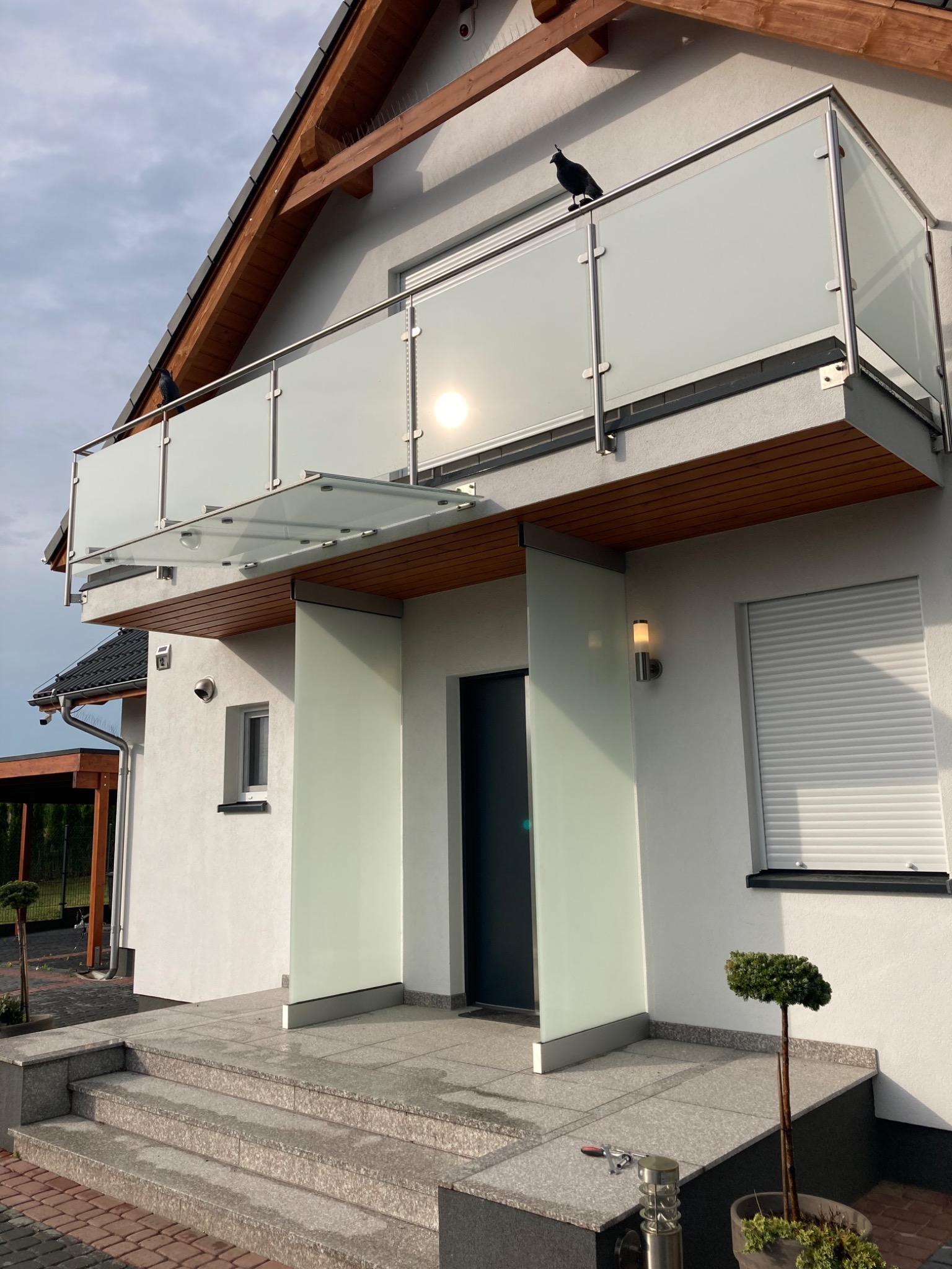 Nowoczesny dom z balkonem zabezpieczonym szklaną balustradą ze stalowymi słupkami, na której siedzi czarny ptak. Wejście do domu osłonięte szklanym daszkiem i ściankami, z granitowymi schodami.