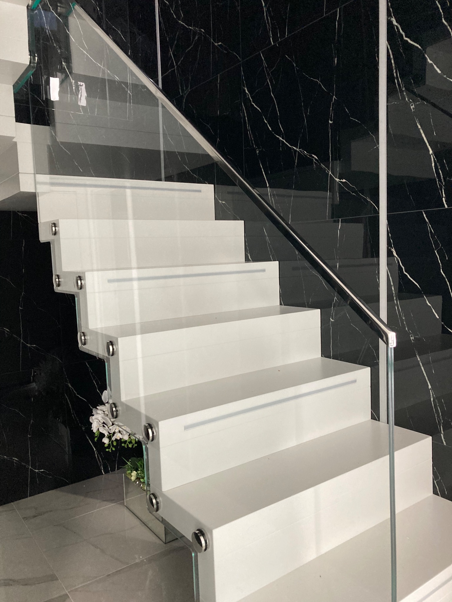 Nowoczesne, białe schody z przezroczystą balustradą szklaną i chromowanymi detalami, na tle ściany z czarnego marmuru z białymi żyłkami. Dekoracja kwiatowa u podstawy schodów.