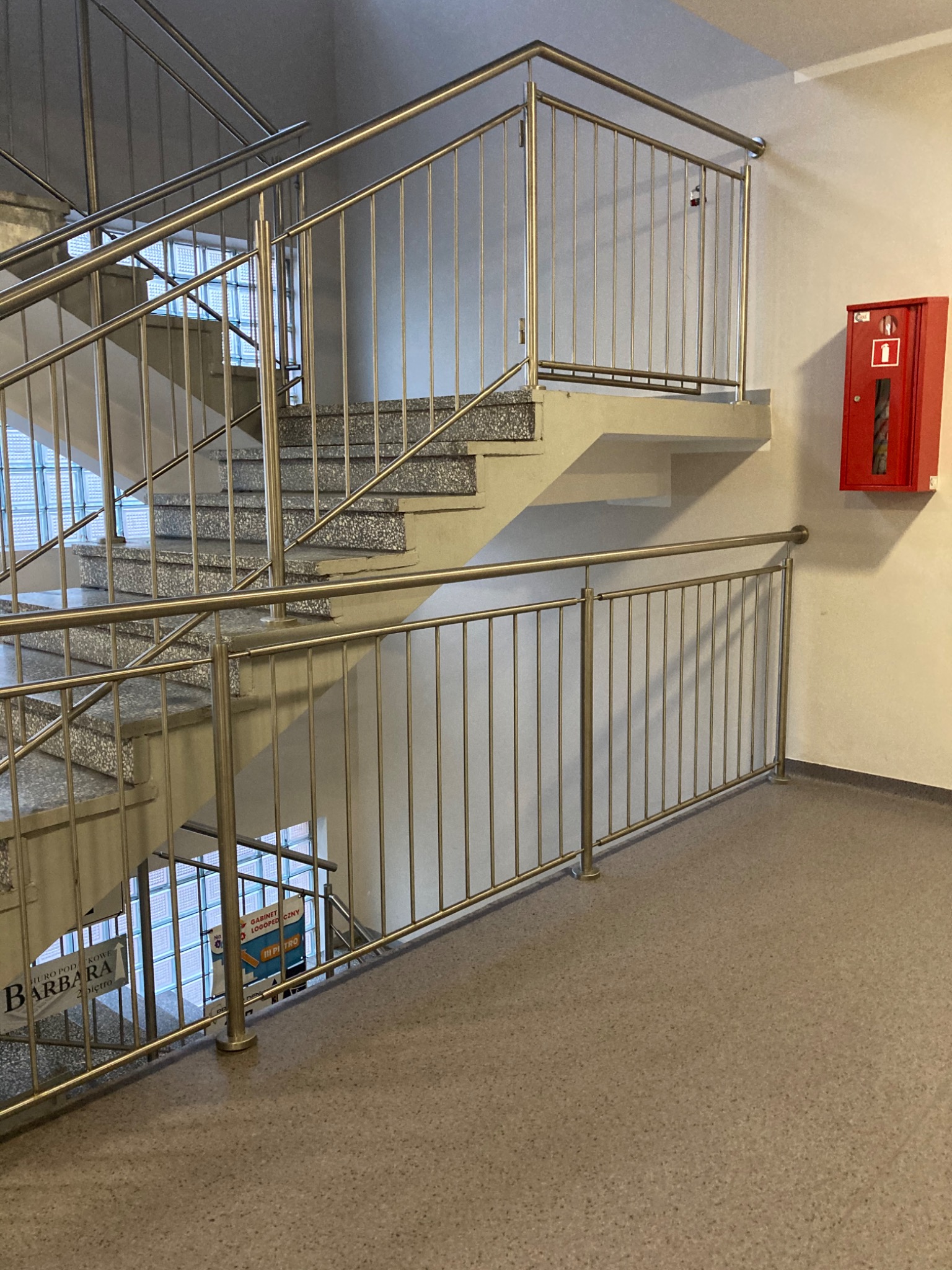 Klatka schodowa w budynku użyteczności publicznej z betonowymi schodami i metalową balustradą, widoczna skrzynka przeciwpożarowa na ścianie.