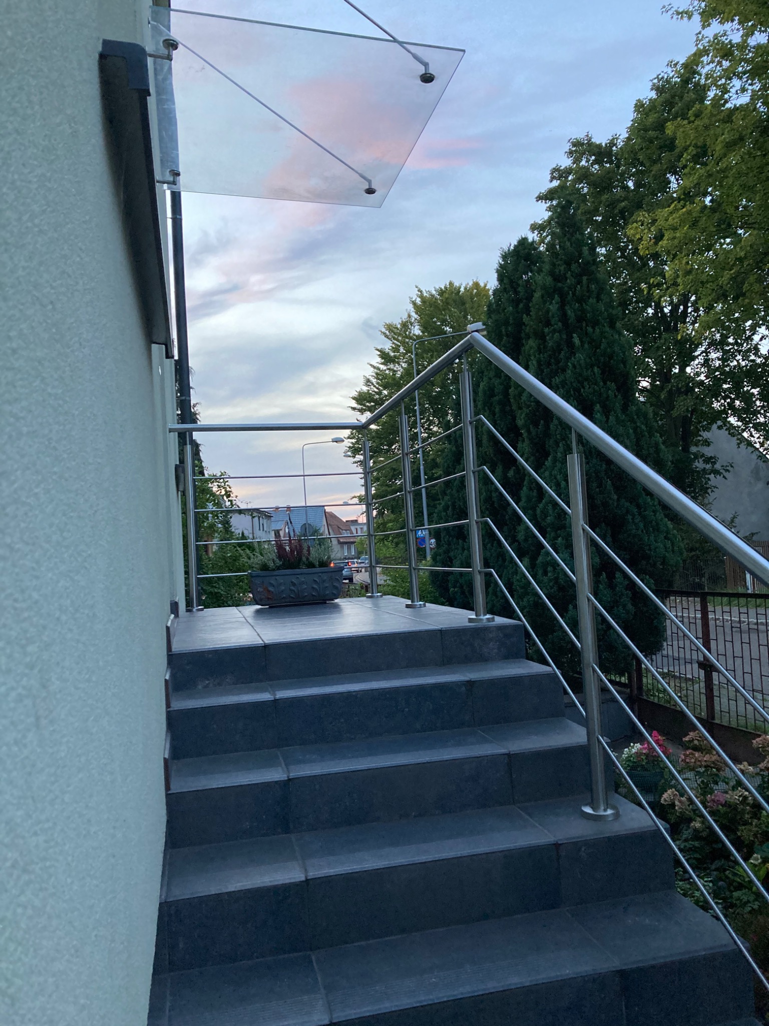 Zewnętrzne schody z ciemnych płytek prowadzące do wejścia z balustradą ze stali nierdzewnej i szklanym zadaszeniem, widok na okolicę w tle.