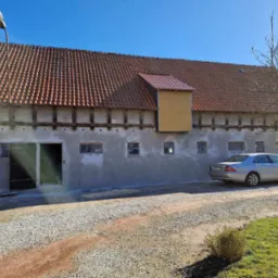 Odnowiony budynek gospodarczy z nowym tynkiem, widoczny srebrny samochód osobowy i czerwony traktor z przodu.