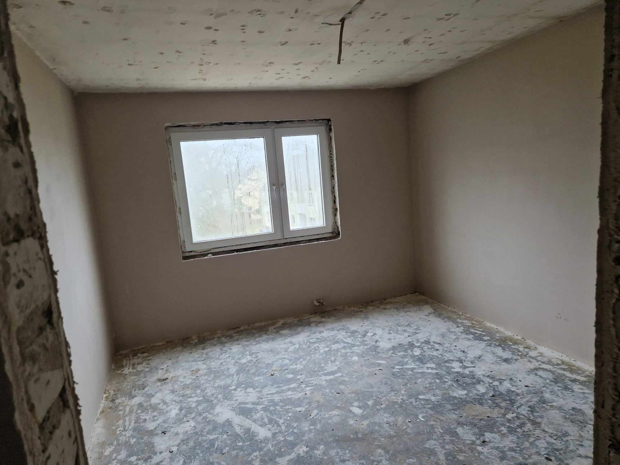 Surowe wnętrze pokoju po tynkowaniu, z widocznym oknem i niedokończoną podłogą, ukazujące etap wykończenia wnętrza.