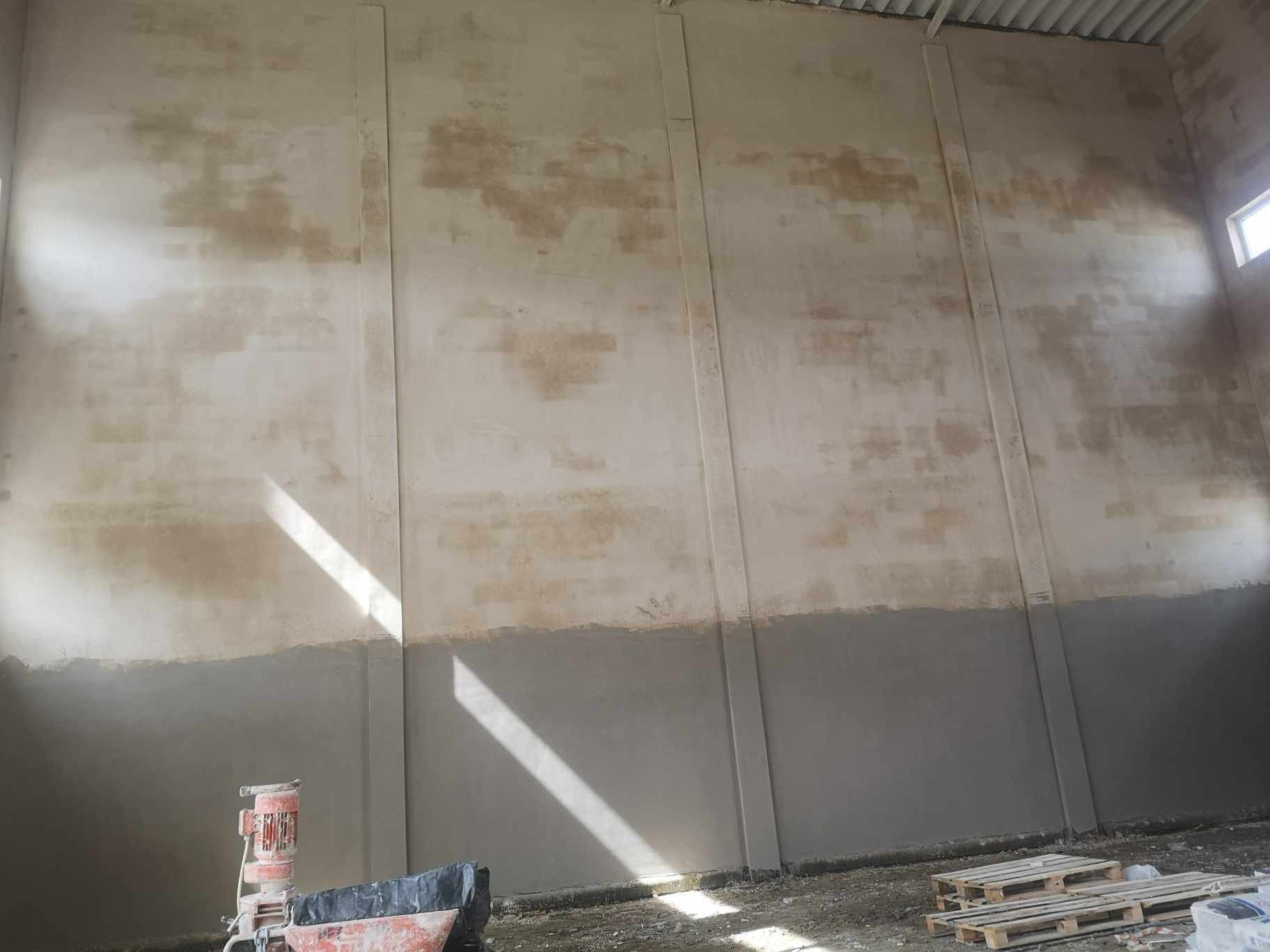 Ściana w hali przemysłowej częściowo pokryta świeżym tynkiem, z widocznym pasmem starego tynku na górze, maszyna do nakładania tynku w lewym dolnym rogu, oraz jasny promień światła słonecznego...