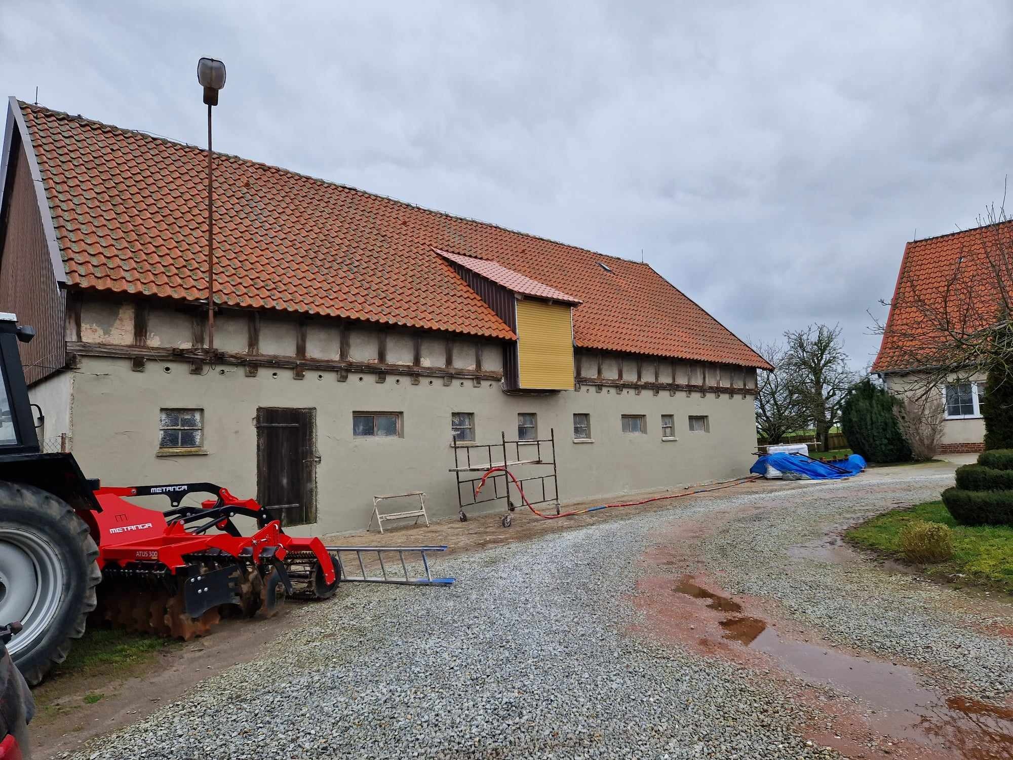 Stary budynek gospodarczy z czerwoną dachówką, świeżo otynkowany, z widocznym sprzętem rolniczym i rusztowaniem, przygotowany do dalszych prac wykończeniowych.