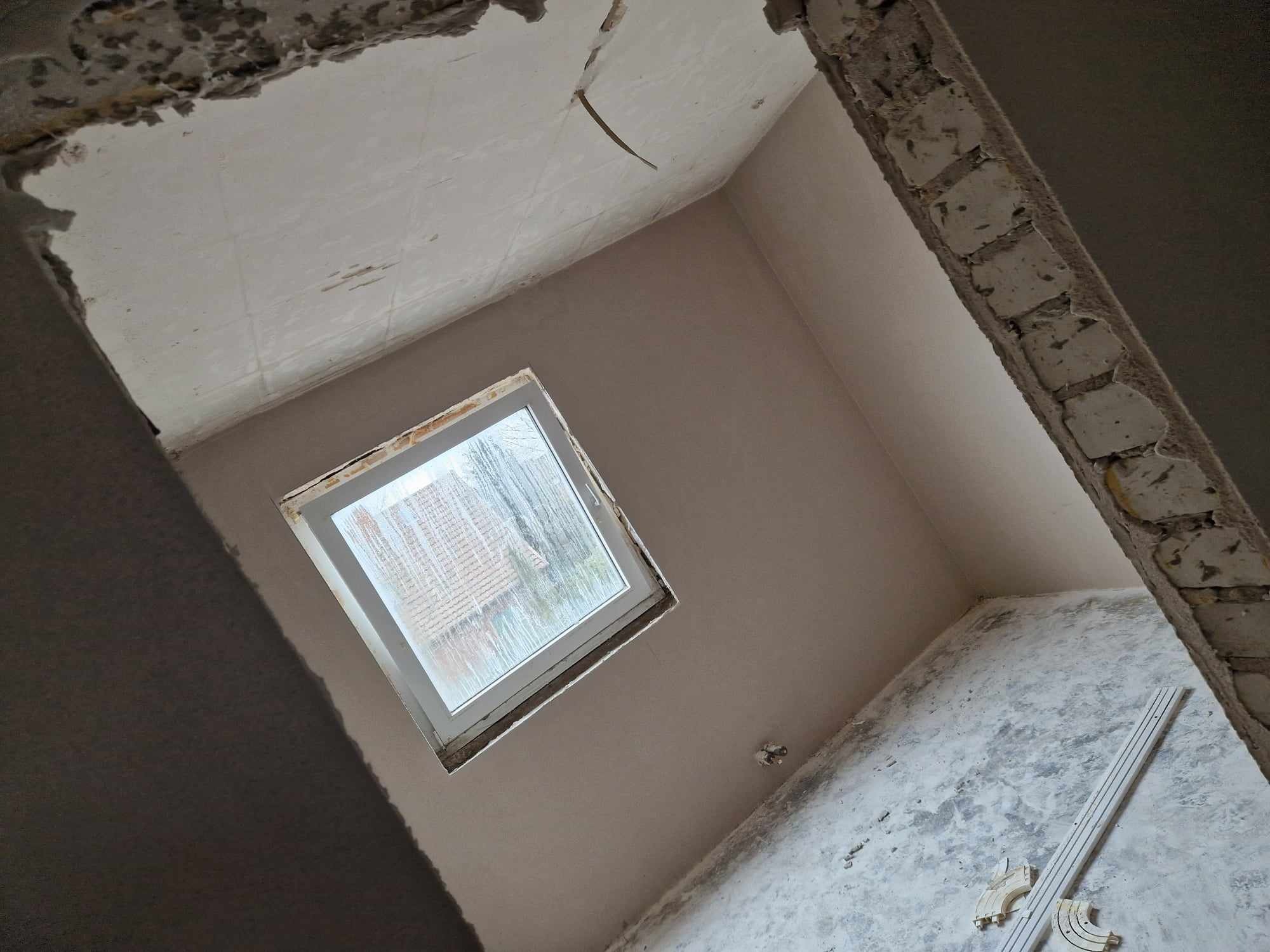Świeżo otynkowane wnętrze z oknem, widoczne niedoskonałości sufitu i resztki zaprawy na podłodze, listwa przypodłogowa leżąca na podłodze.