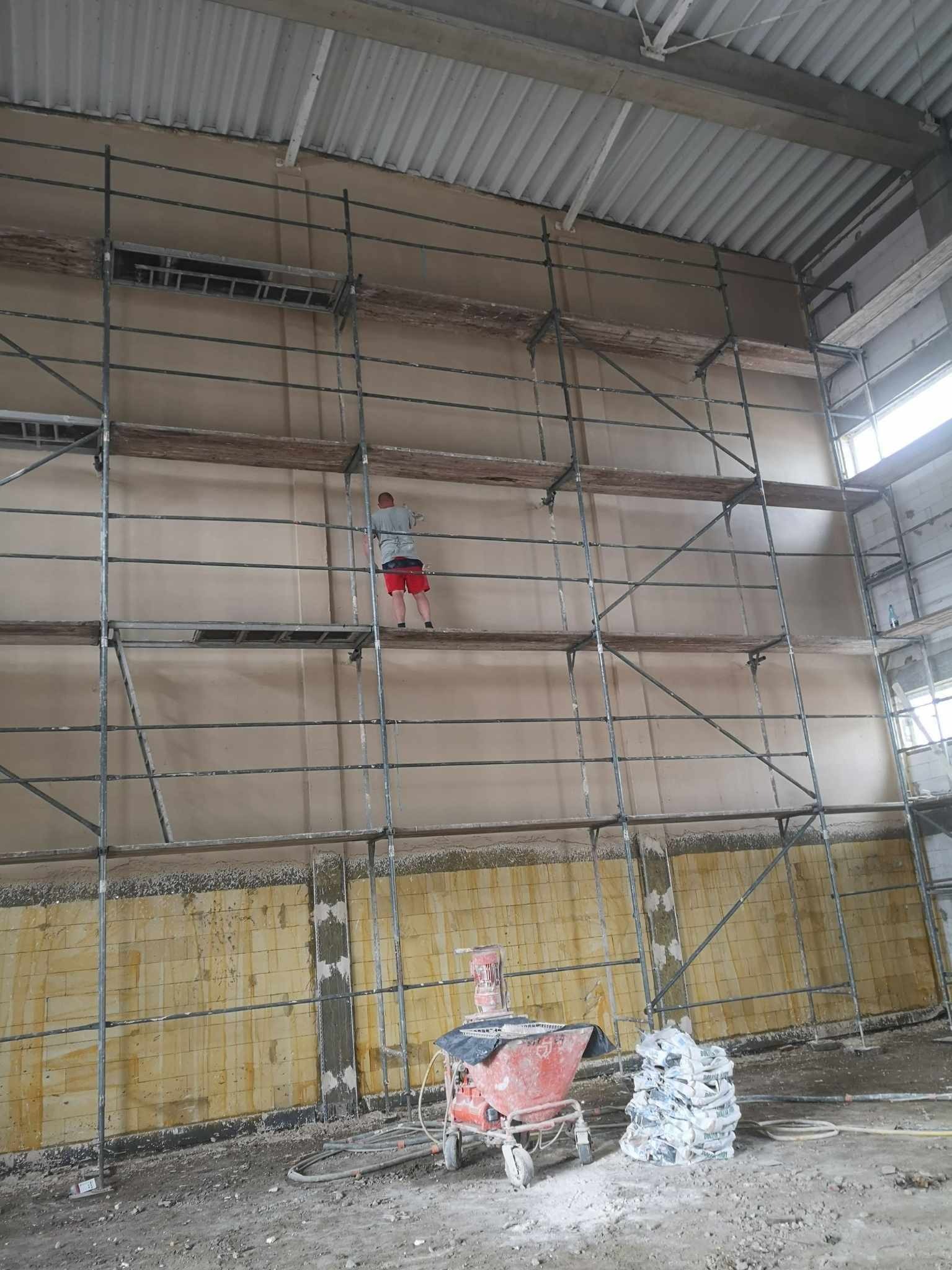 Pracownik na rusztowaniu aplikuje tynk na ścianę w hali przemysłowej, widoczna maszyna do tynkowania i worki z zaprawą.