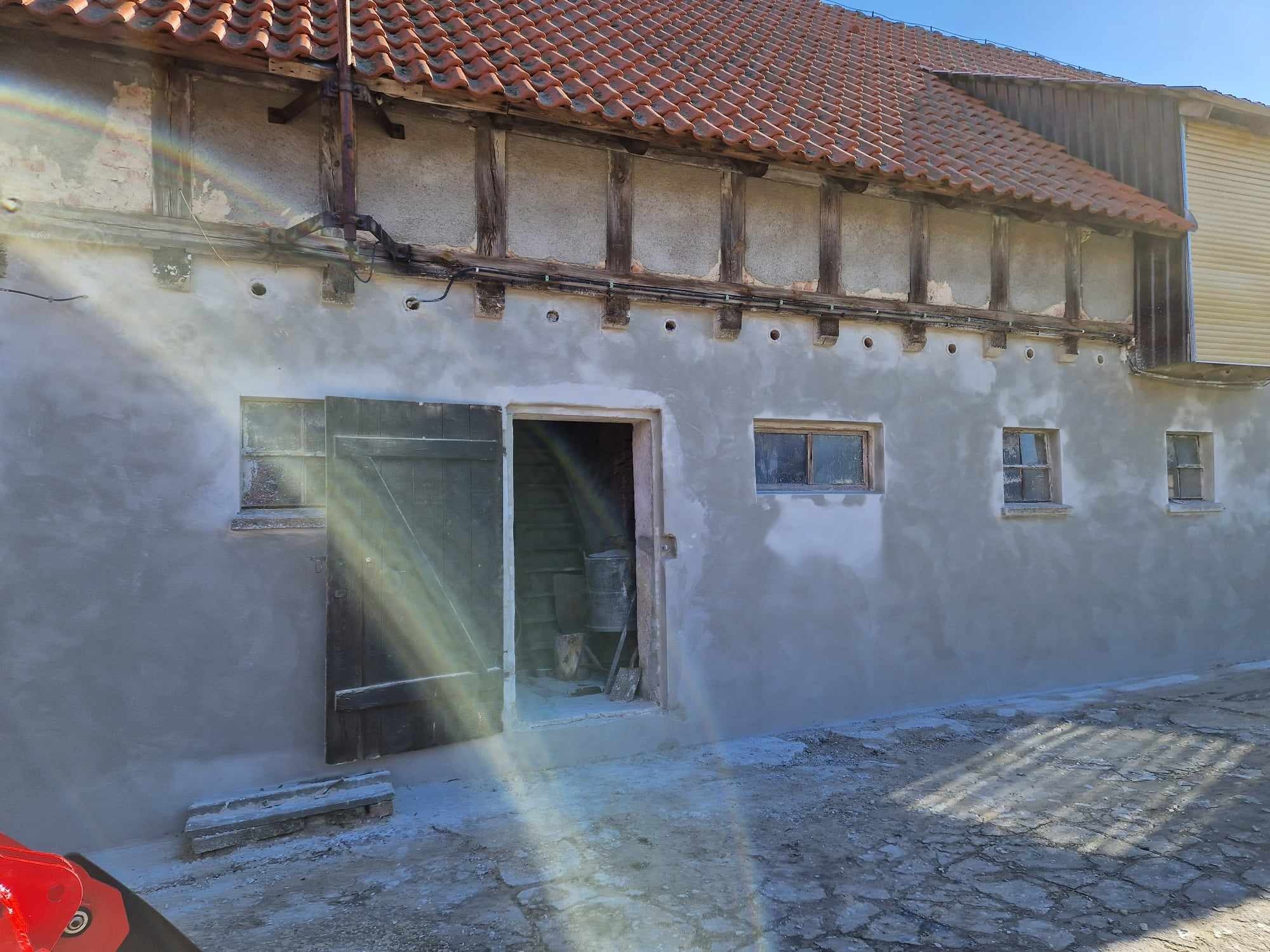 Budynek gospodarczy po nałożeniu szarego tynku maszynowego, widoczne drewniane elementy konstrukcyjne, czerwona dachówka, otwarte drzwi do wnętrza z widocznymi narzędziami.