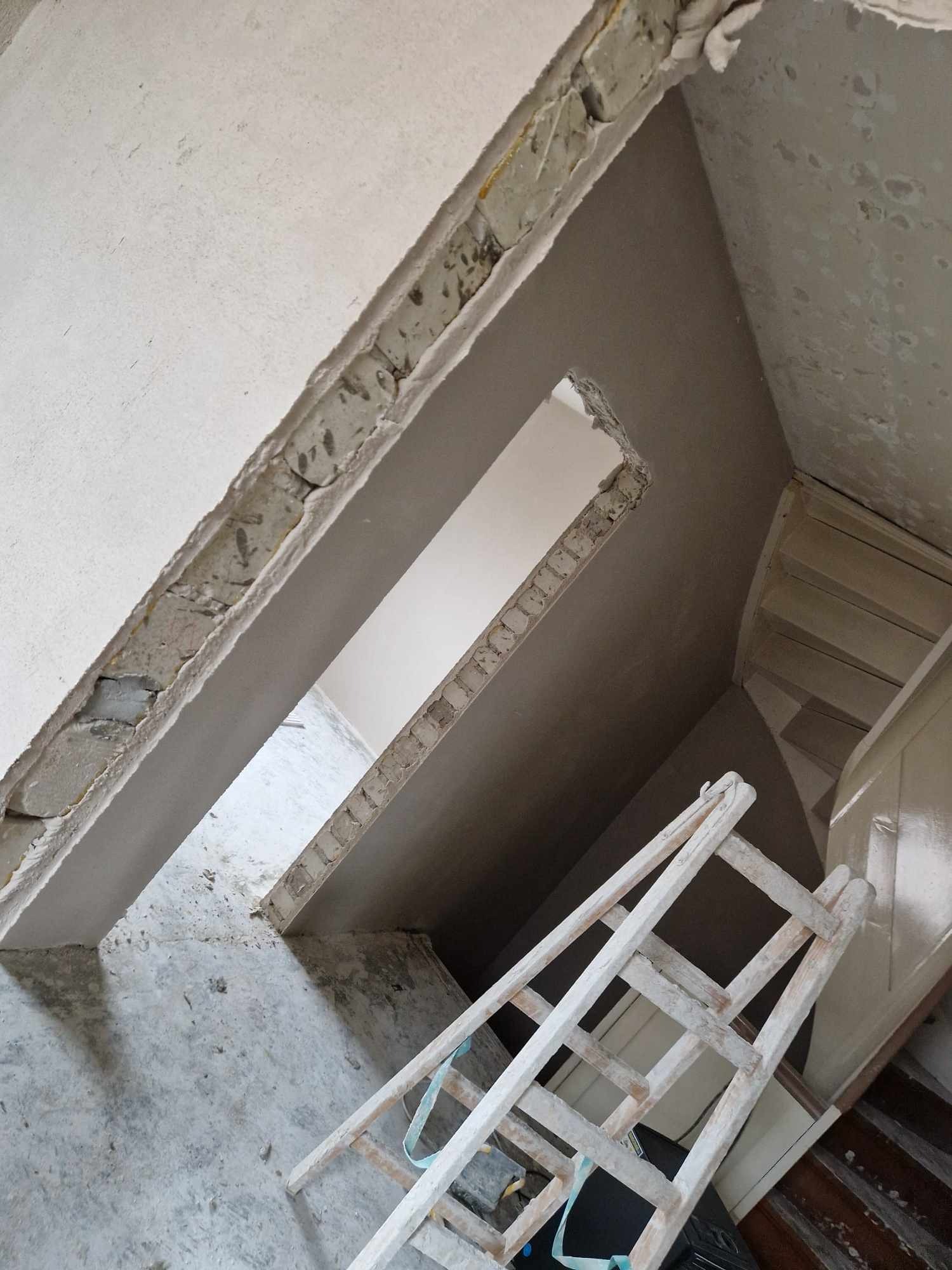 Świeżo nałożona, gładka, szara powłoka na ścianach i suficie, widoczne fragmenty cegieł w otworach, biała, drewniana drabina oparta o schody, surowy beton na podłodze.