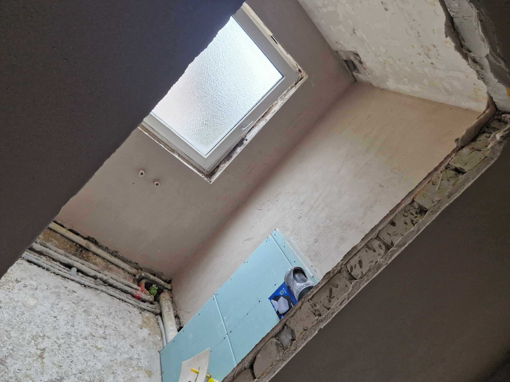 Wnętrze w trakcie remontu: widok na okno dachowe, surowe ściany, odsłonięte rury i płyty gipsowo-kartonowe przed nałożeniem gładzi.