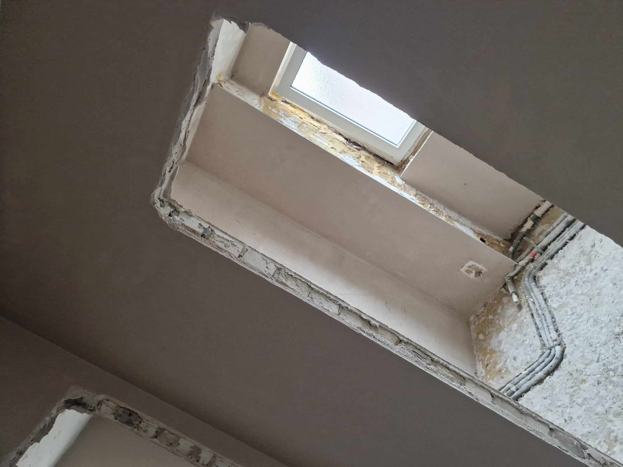 Surowy stan sufitu po nałożeniu wstępnej warstwy tynku w okolicy okna dachowego; widoczne przewody elektryczne i niedoskonałości wykończenia.