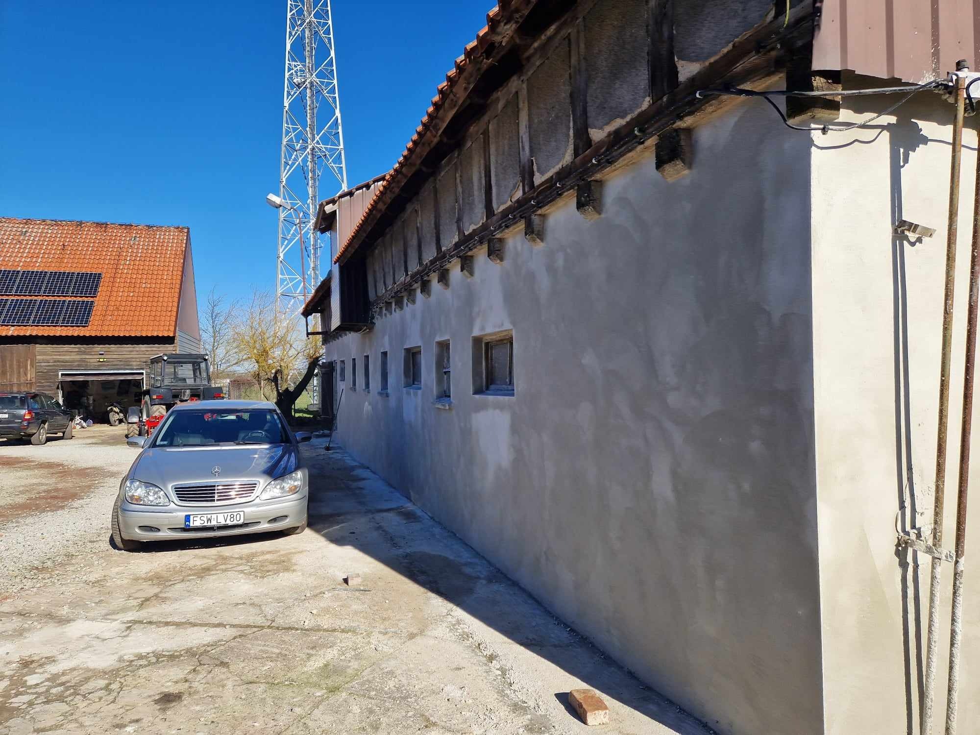 Świeżo otynkowana ściana budynku gospodarczego z widocznymi drewnianymi belkami i instalacjami, na tle wieży telekomunikacyjnej i samochodu osobowego.