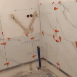 Białe, marmurowe płytki na ścianie łazienki w trakcie montażu. Widoczne krzyżyki dystansowe, poziomice laserowe i fragment instalacji wodnej. Etap prac glazurniczych.