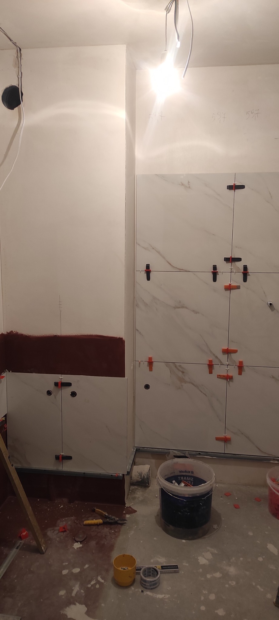 Ściana w trakcie układania płytek imitujących marmur, z użyciem krzyżyków dystansowych i poziomicy, widać przygotowane podłoże. Prace w łazience.