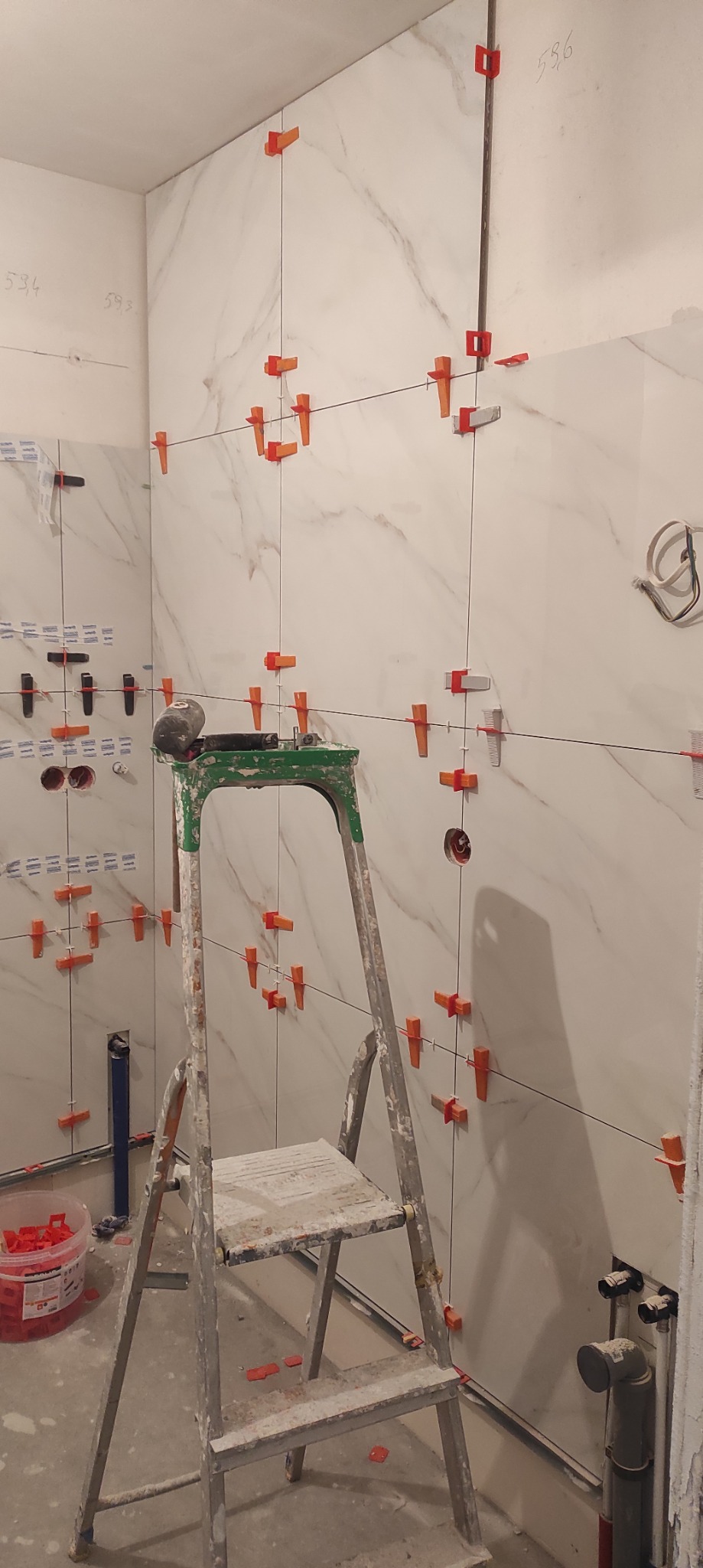 Białe płytki imitujące marmur na ścianie łazienki, z systemem poziomowania płytek. Widoczna drabina malarska i narzędzia glazurnicze. W tle instalacja wodno-kanalizacyjna.