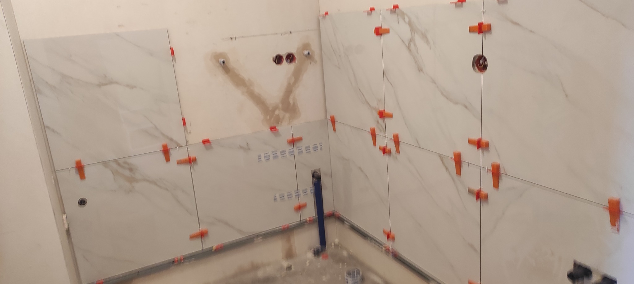 Białe, marmurowe płytki na ścianie łazienki w trakcie montażu. Widoczne krzyżyki dystansowe, poziomice laserowe i fragment instalacji wodnej. Etap prac glazurniczych.