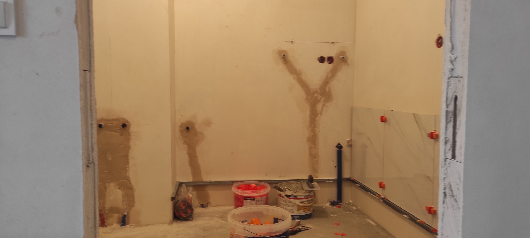 Łazienka w trakcie remontu: częściowo ułożone białe płytki z czerwonymi krzyżykami dystansowymi, przygotowane ściany z instalacjami i wiaderka z zaprawą na podłodze.