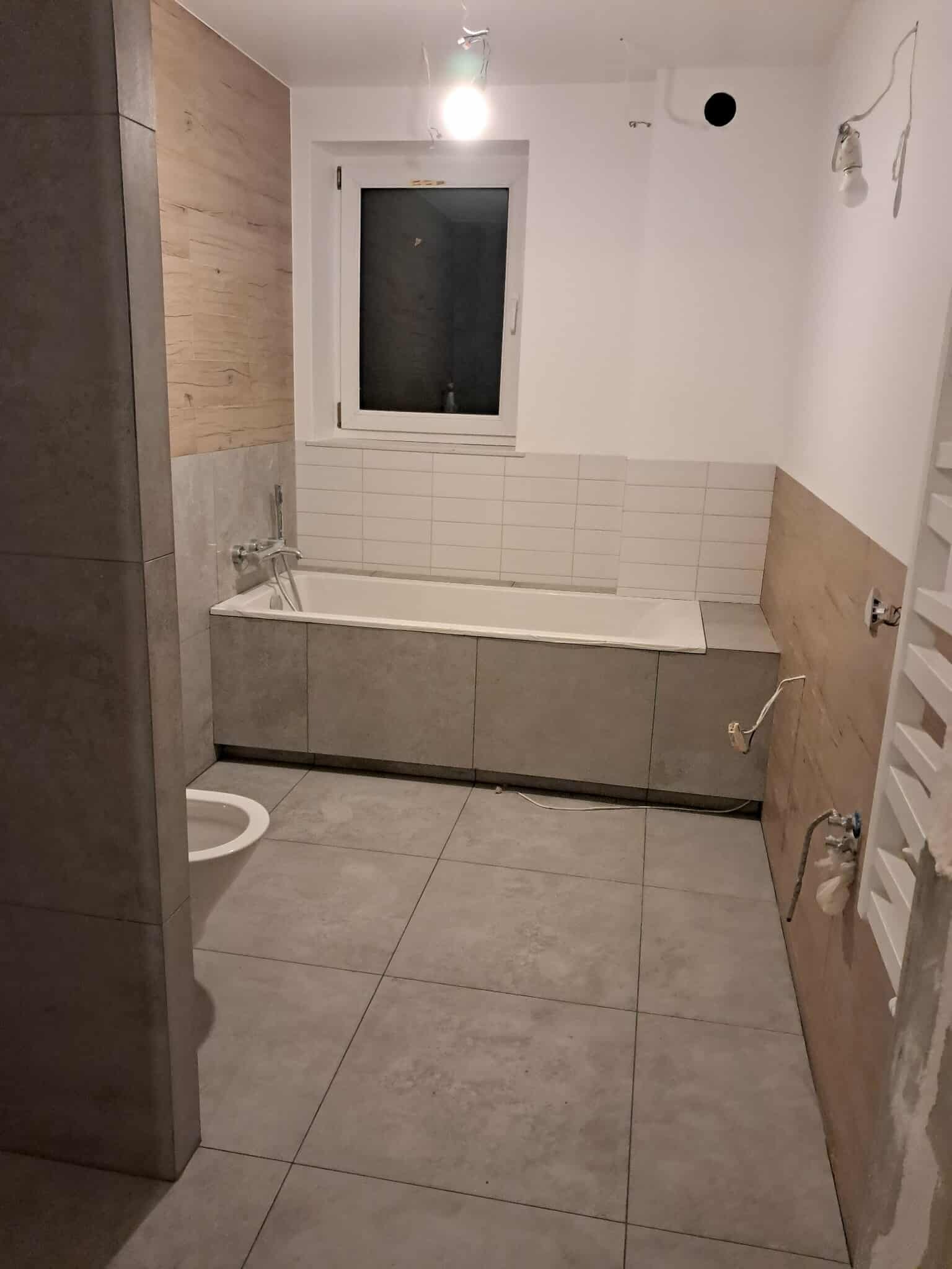 Nowoczesna łazienka w trakcie wykańczania: szare płytki, zabudowana wanna, drewnopodobne panele ścienne. Widoczne niedokończone instalacje.