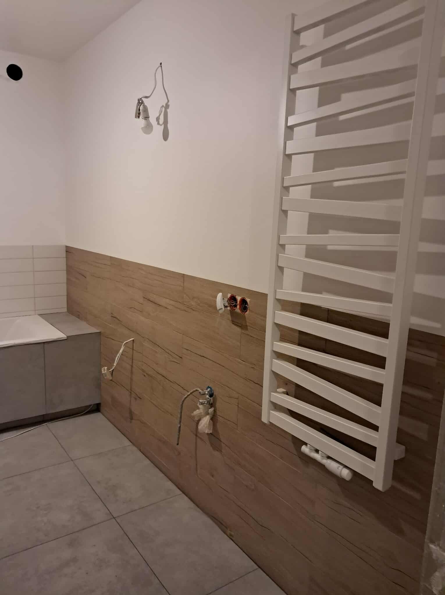 Remontowana łazienka z grzejnikiem drabinkowym, częściowo wykończona drewnopodobną okładziną ścienną i szarą podłogą. Widoczne niedokończone instalacje.