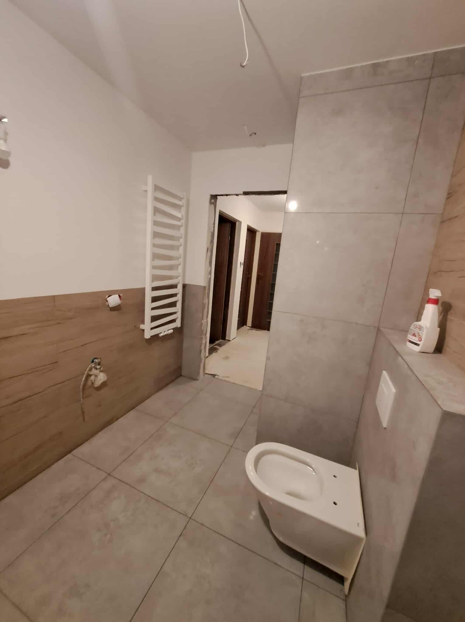 Jasna łazienka w trakcie remontu: szare płytki, podwieszana toaleta, grzejnik drabinkowy. Widoczne niedokończone prace i akcesoria. Ujęcie z perspektywy wejścia.