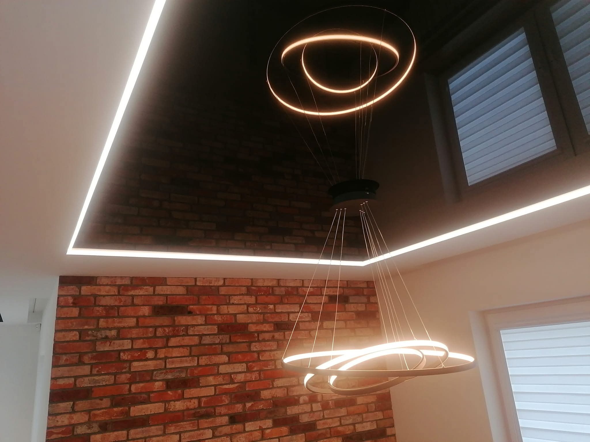 Nowoczesne oświetlenie LED w suficie podwieszanym, z ceglaną ścianą i designerską lampą wiszącą. Minimalistyczny design wnętrza z akcentem na detale świetlne.