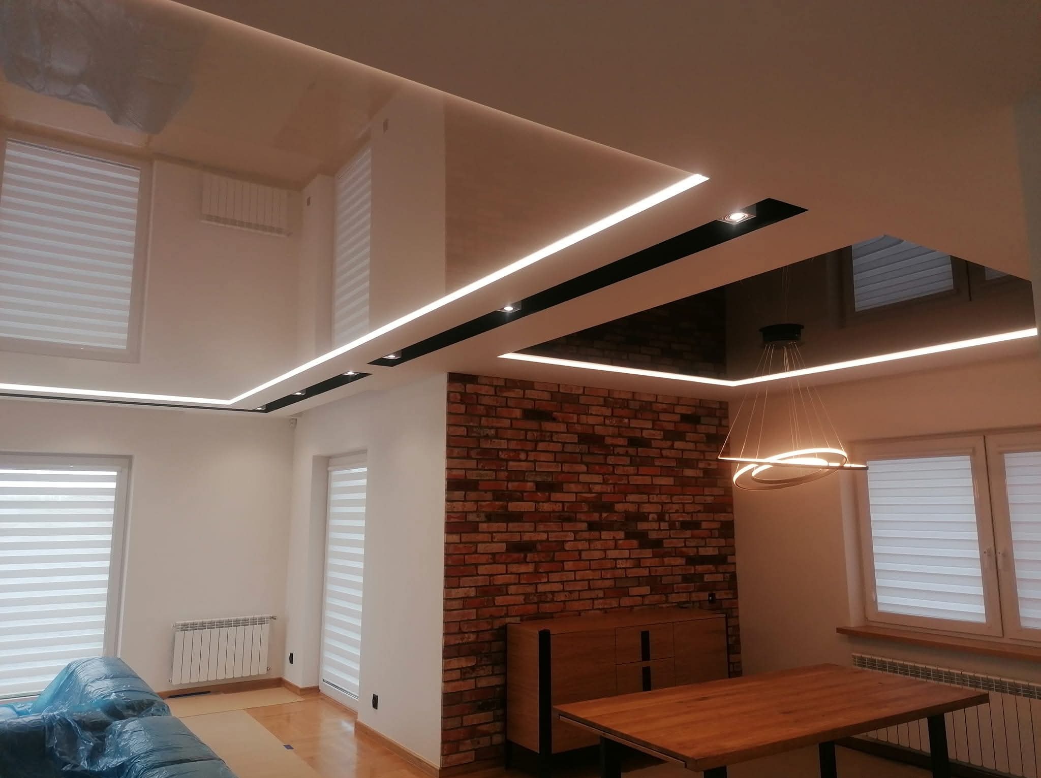 Nowoczesne wnętrze z podwieszanym sufitem, oświetleniem LED i ceglaną ścianą. Widoczne okna z żaluzjami, drewniany stół i komoda. Częściowo zakryty mebel folią ochronną.
